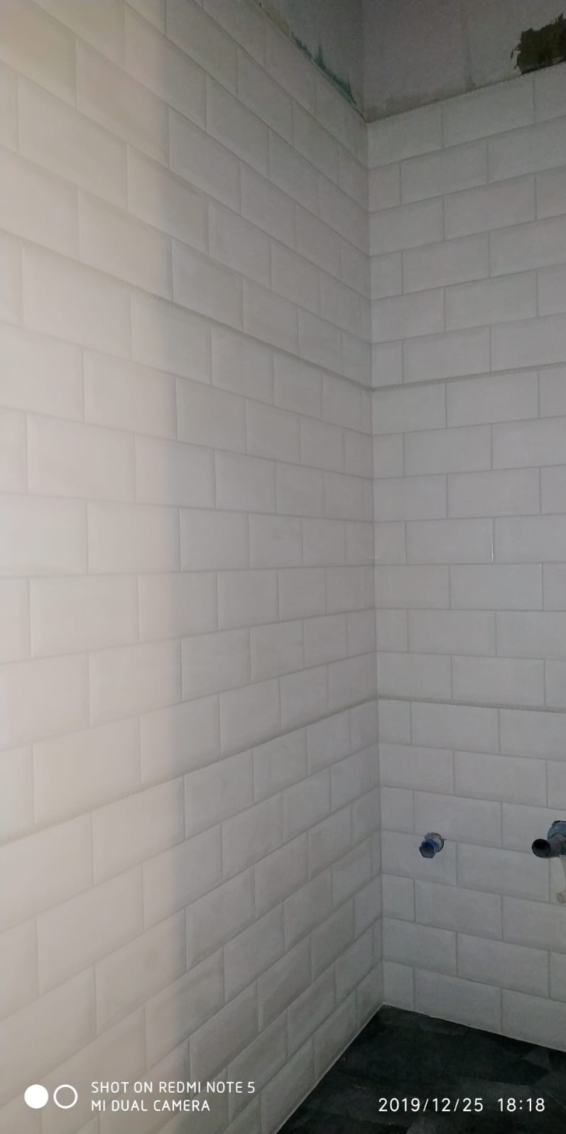 Białe, prostokątne płytki ułożone w cegiełkę na ścianach w rogu łazienki. Widoczne fragmenty instalacji wodnej i niedokończone wykończenie sufitu.