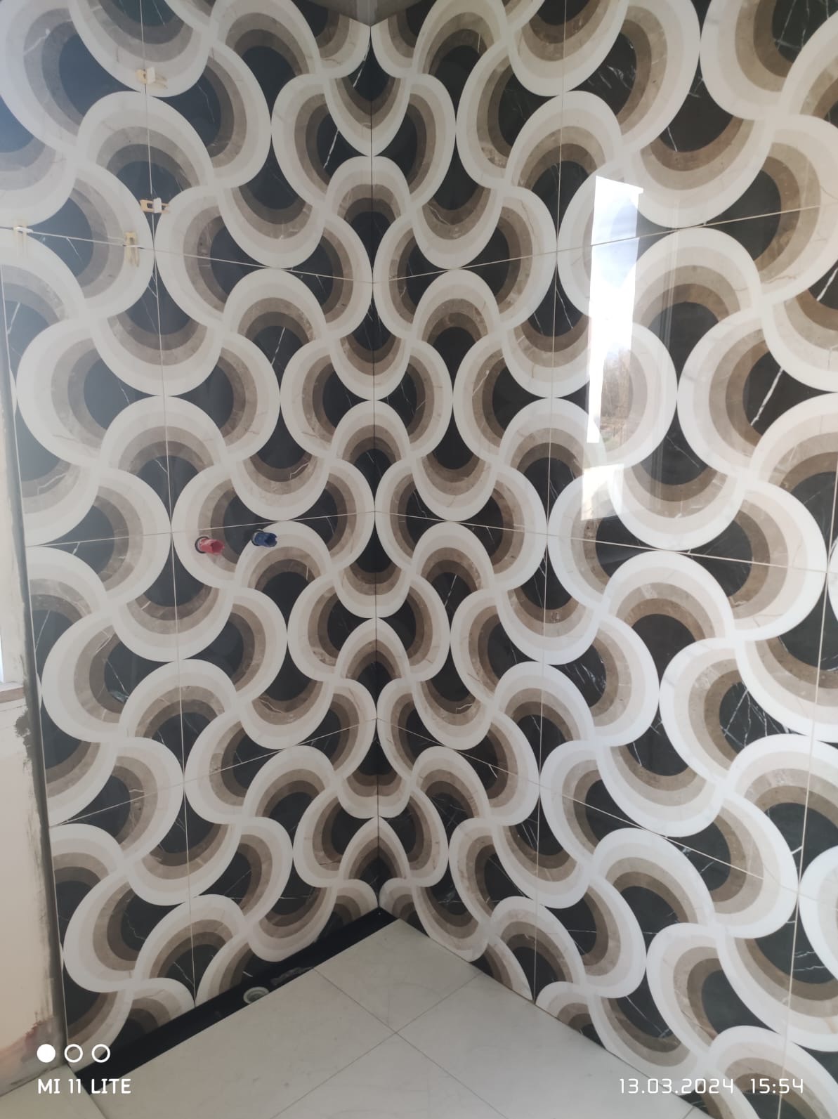 Ściana wyłożona błyszczącymi, wzorzystymi płytkami w odcieniach bieli, beżu i czerni, tworzącymi geometryczny, koncentryczny deseń. Widoczne fugi i fragment podłogi.