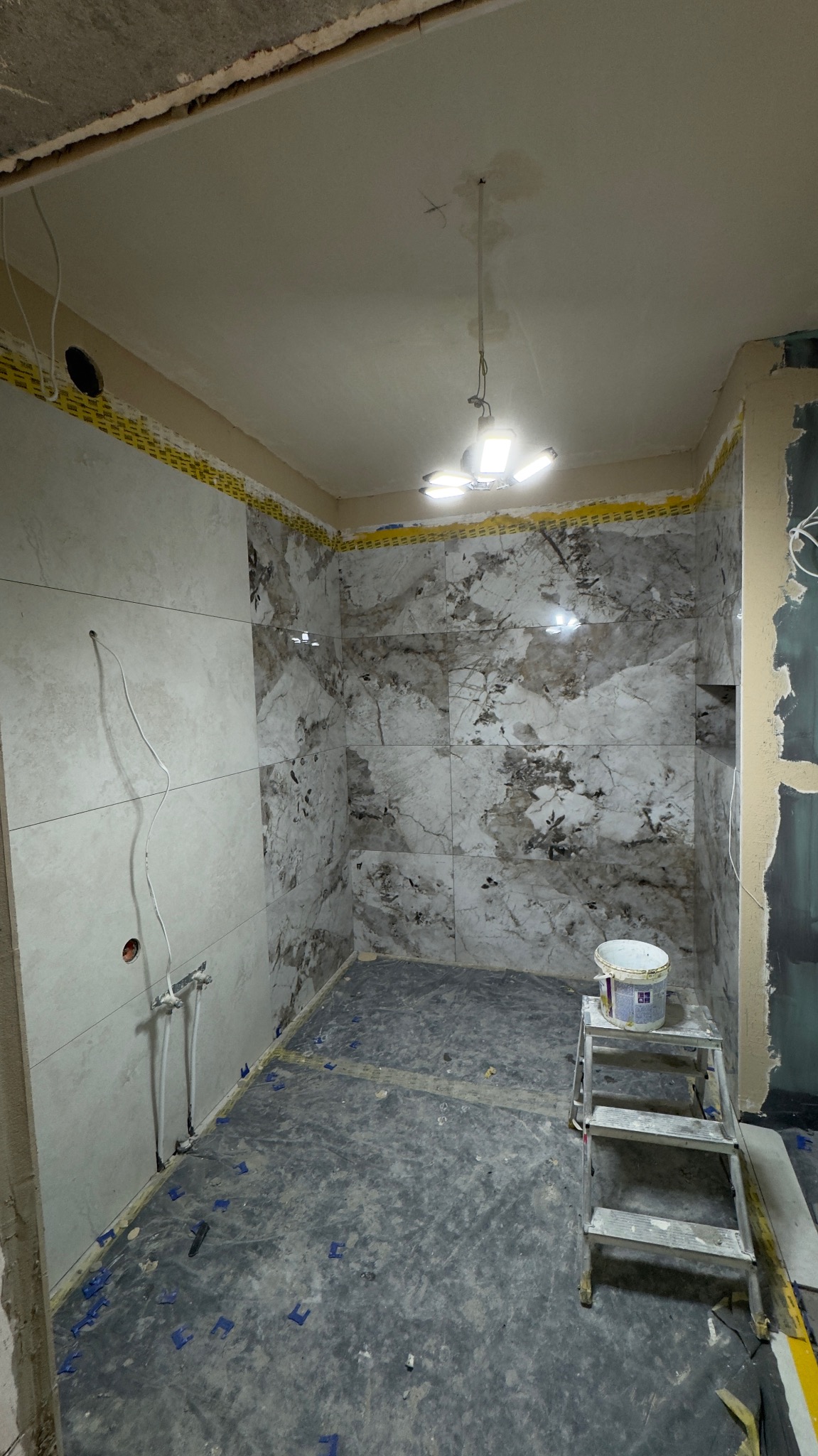 Wnętrze łazienki w trakcie prac wykończeniowych: ułożone płytki imitujące marmur na ścianach, widoczne narzędzia glazurnicze i drabina z wiadrem farby.