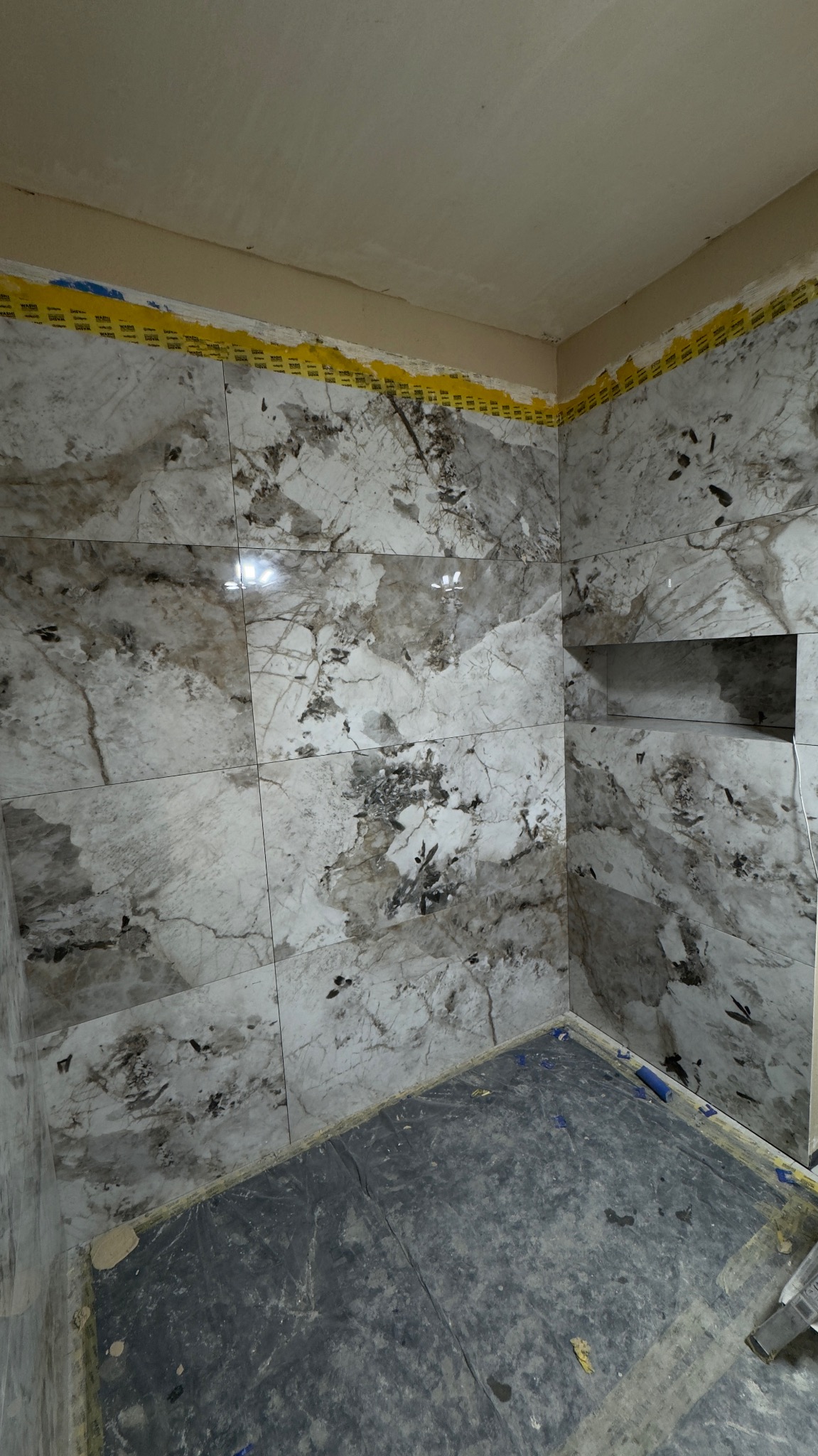 Wykończona łazienka z marmurowymi płytkami i wnęką. Zabezpieczona folia na podłodze i taśma malarska przy suficie. Widok na narożnik i fragment podłogi.