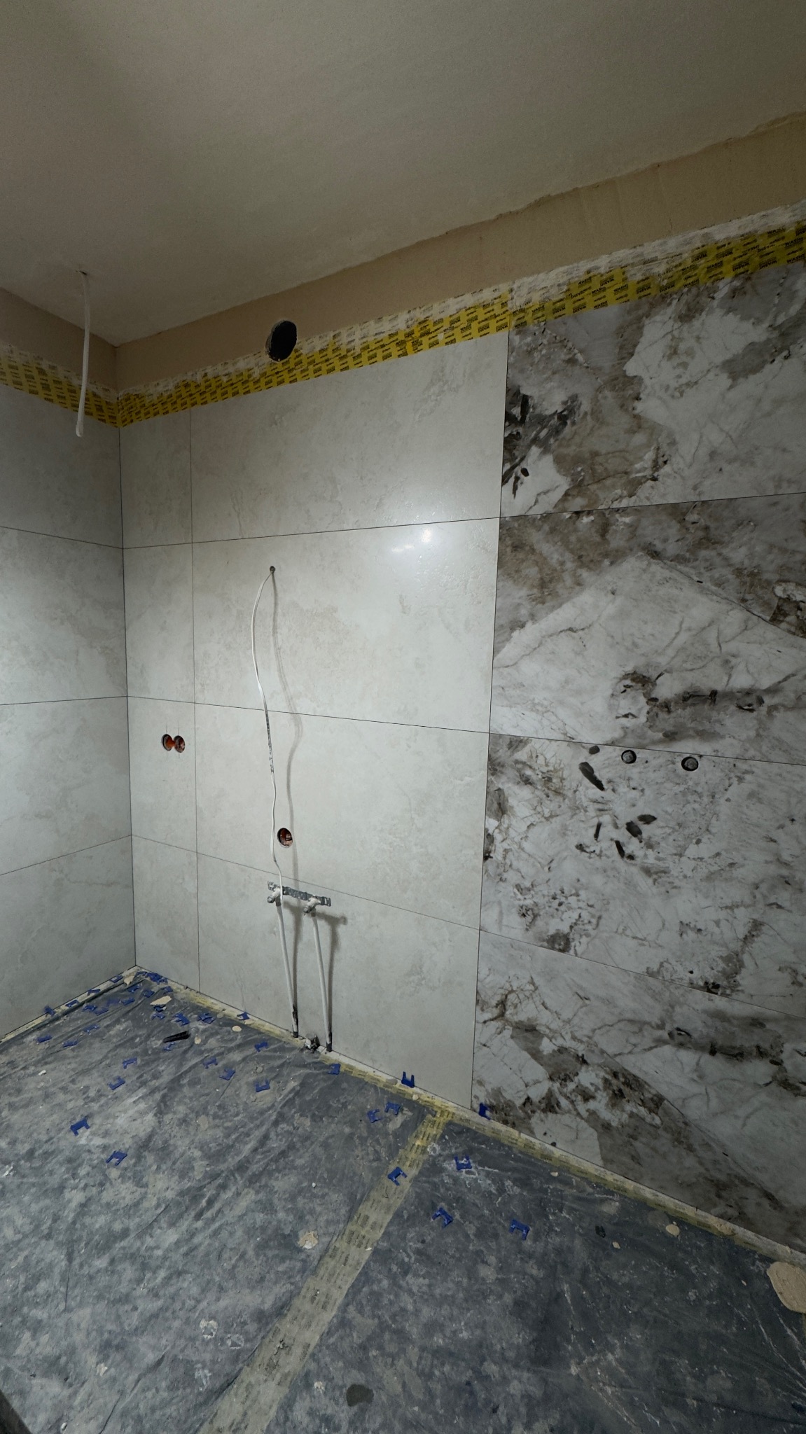 Wnętrze łazienki w trakcie prac. Częściowo ułożone płytki imitujące marmur na ścianach, widoczne instalacje wodne i elektryczne, zabezpieczona podłoga.