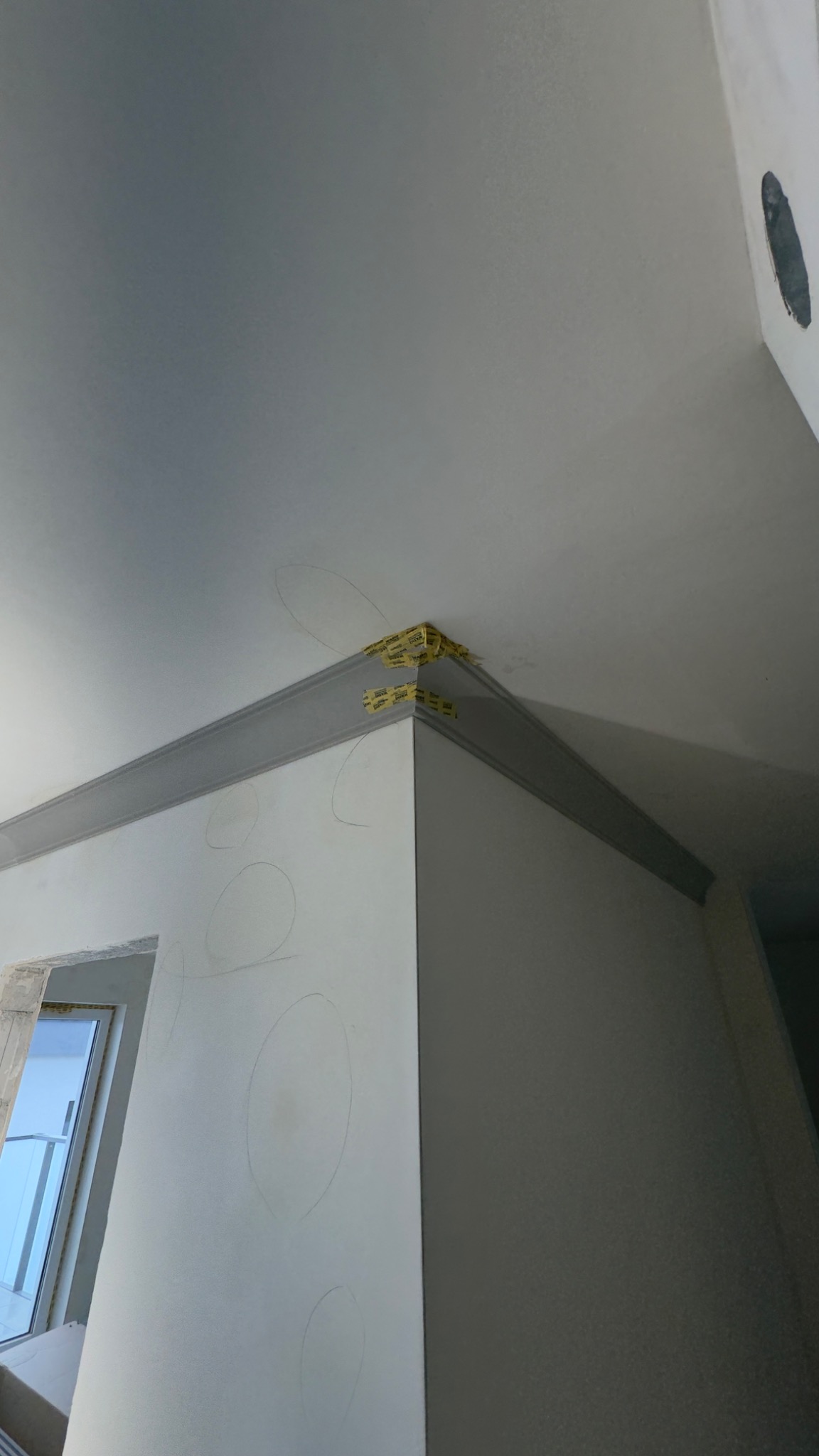 Szara listwa dekoracyjna przy suficie, zabezpieczona taśmą malarską w rogu pomieszczenia. Na ścianie widoczne szkice okręgów, okno w tle.