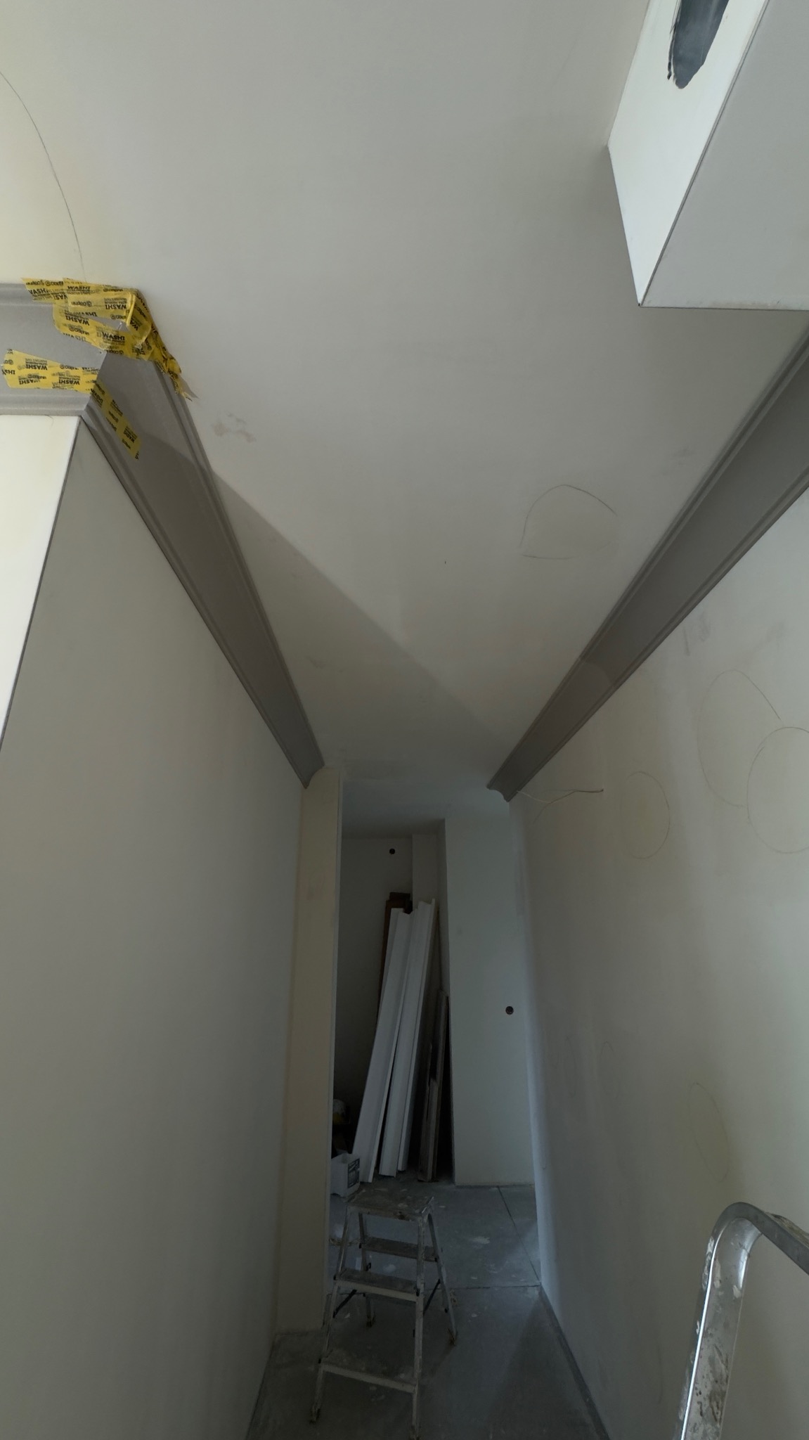 Montaż sztukaterii sufitowej w korytarzu, widoczne taśmy maskujące, drabina i przygotowane listwy. W tle widoczne materiały budowlane. Ujęcie z dołu.