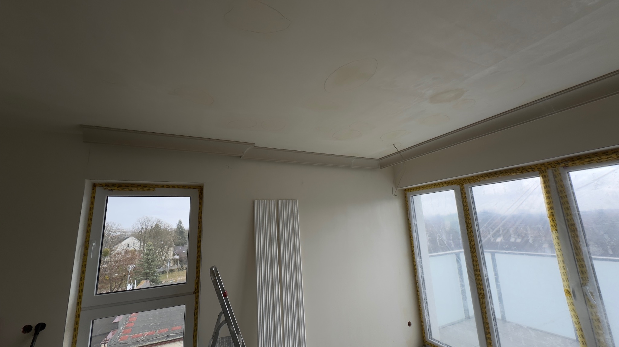 Pokój w trakcie wykańczania: listwy dekoracyjne przy suficie, zabezpieczone okna, ślady po szpachlowaniu na suficie, drabina. Etap prac wykończeniowych.