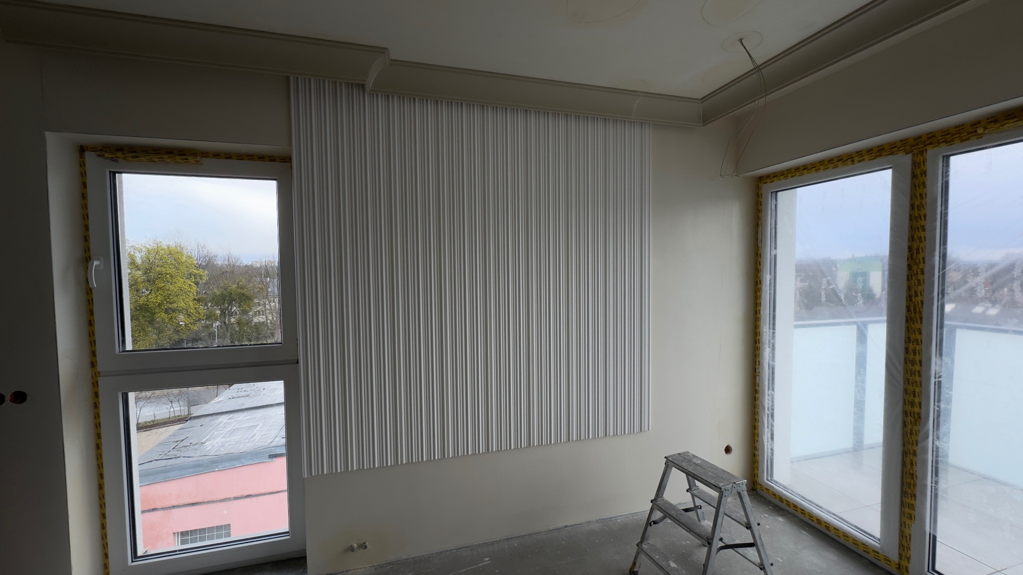 Wykończone wnętrze z dekoracyjną ścianą z paneli 3D, zabezpieczonymi oknami i drabiną. Widoczne detale wykończeniowe sufitu.