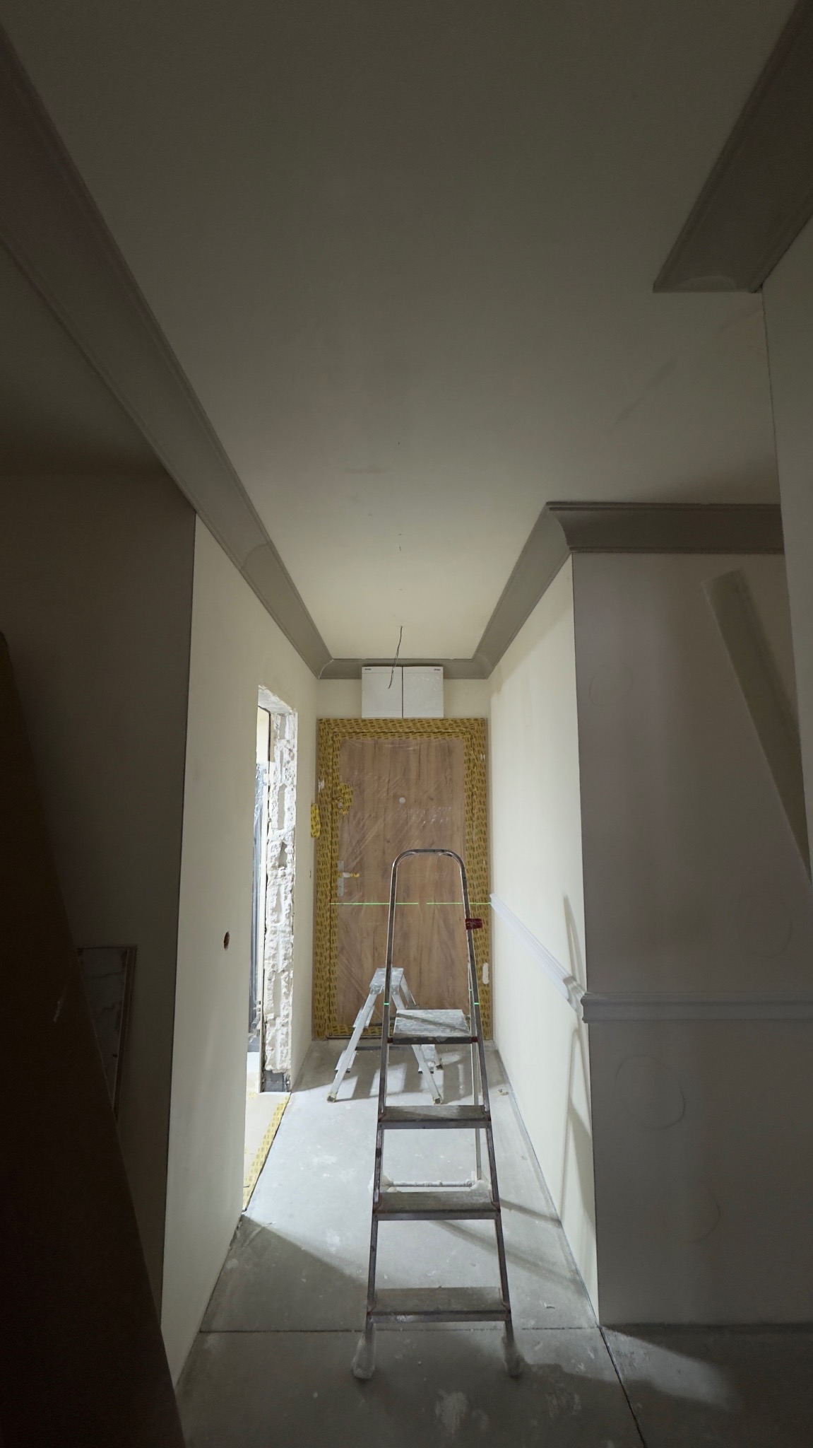Korytarz w trakcie wykańczania: pomalowane ściany, dekoracyjne listwy sufitowe, zabezpieczone drzwi wejściowe. Na podłodze drabina malarska.