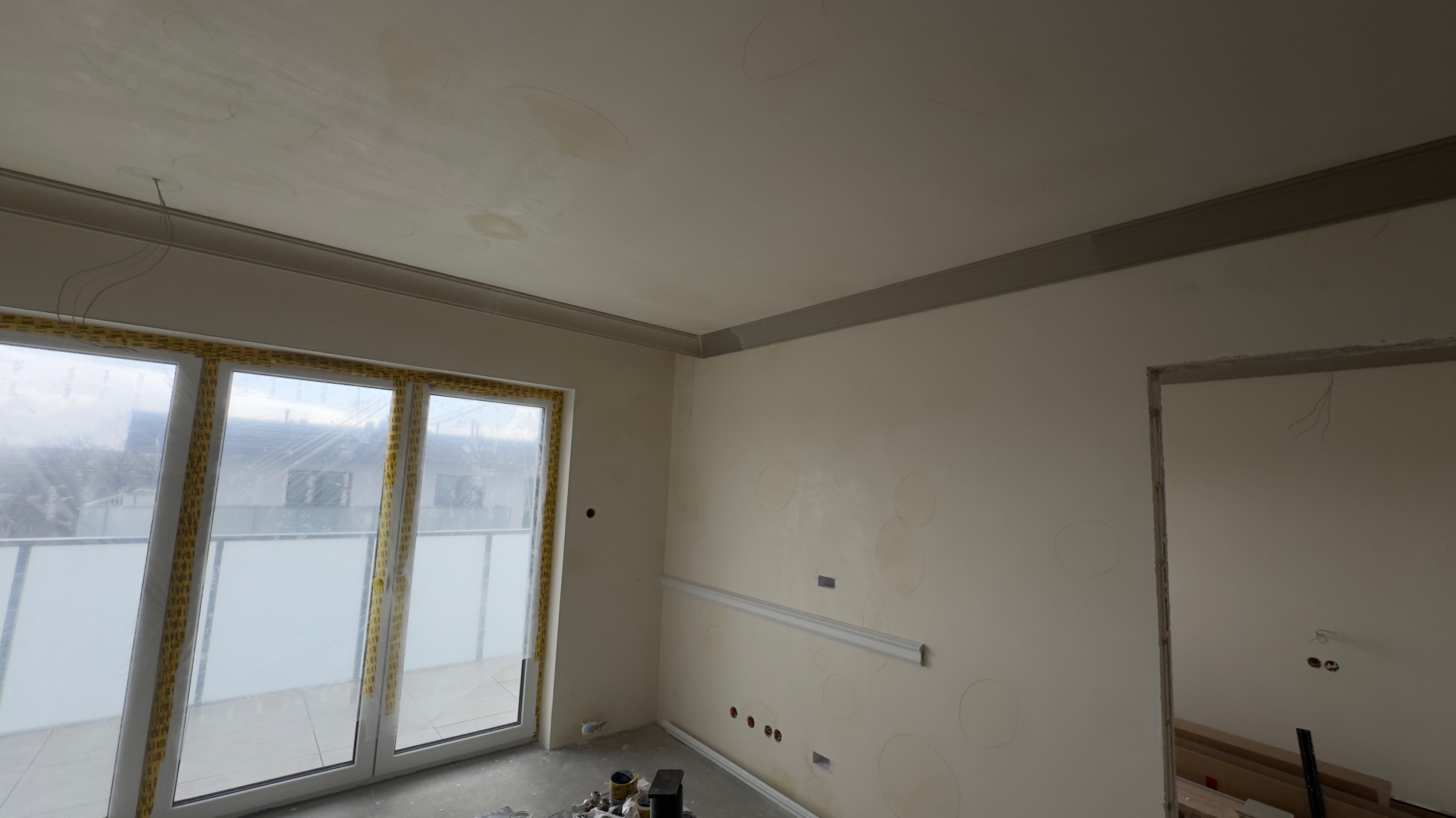 Pokój w trakcie prac wykończeniowych: ściany po szpachlowaniu z widocznymi śladami, listwy dekoracyjne, okno balkonowe zabezpieczone folią, widoczne puszki elektryczne.
