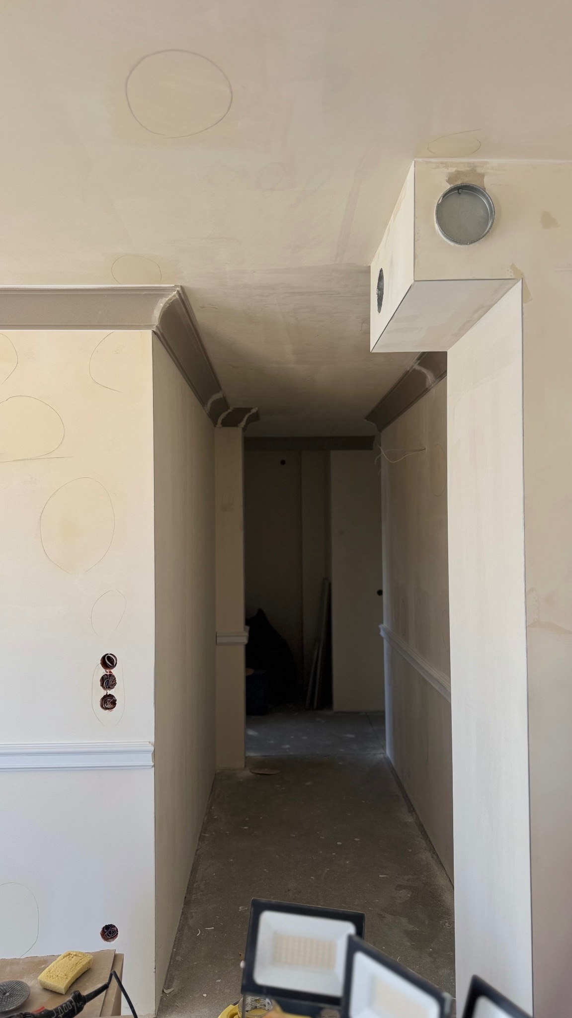 Korytarz w trakcie prac wykończeniowych: ściany po szpachlowaniu, widoczne punkty elektryczne, listwy dekoracyjne, sufit z zaznaczonymi okręgami, lampy robocze na podłodze.