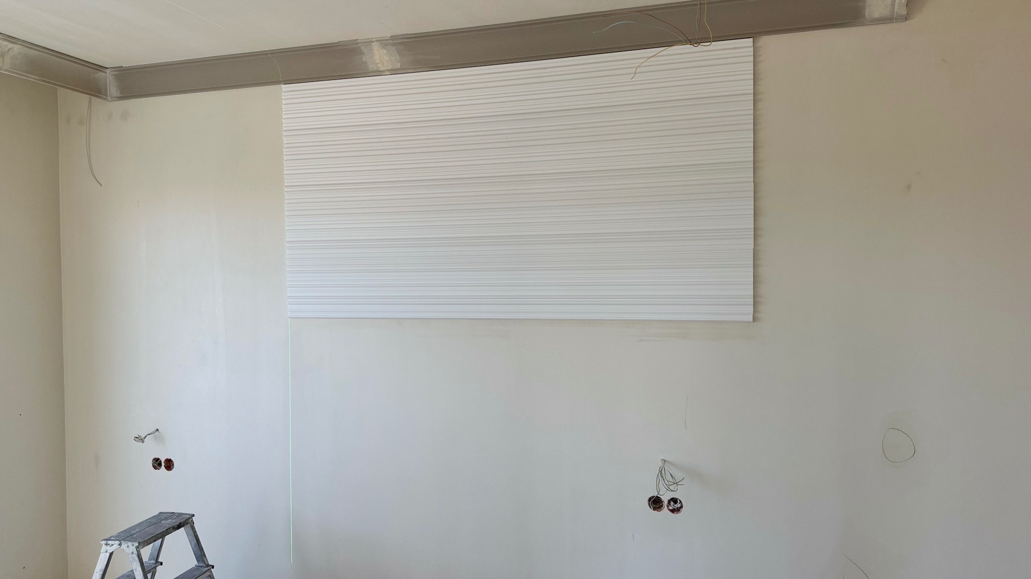 Montaż dekoracyjnej, białej płyty ściennej z poziomymi żłobieniami w jasnym wnętrzu. Widoczna drabina i wystające kable elektryczne.