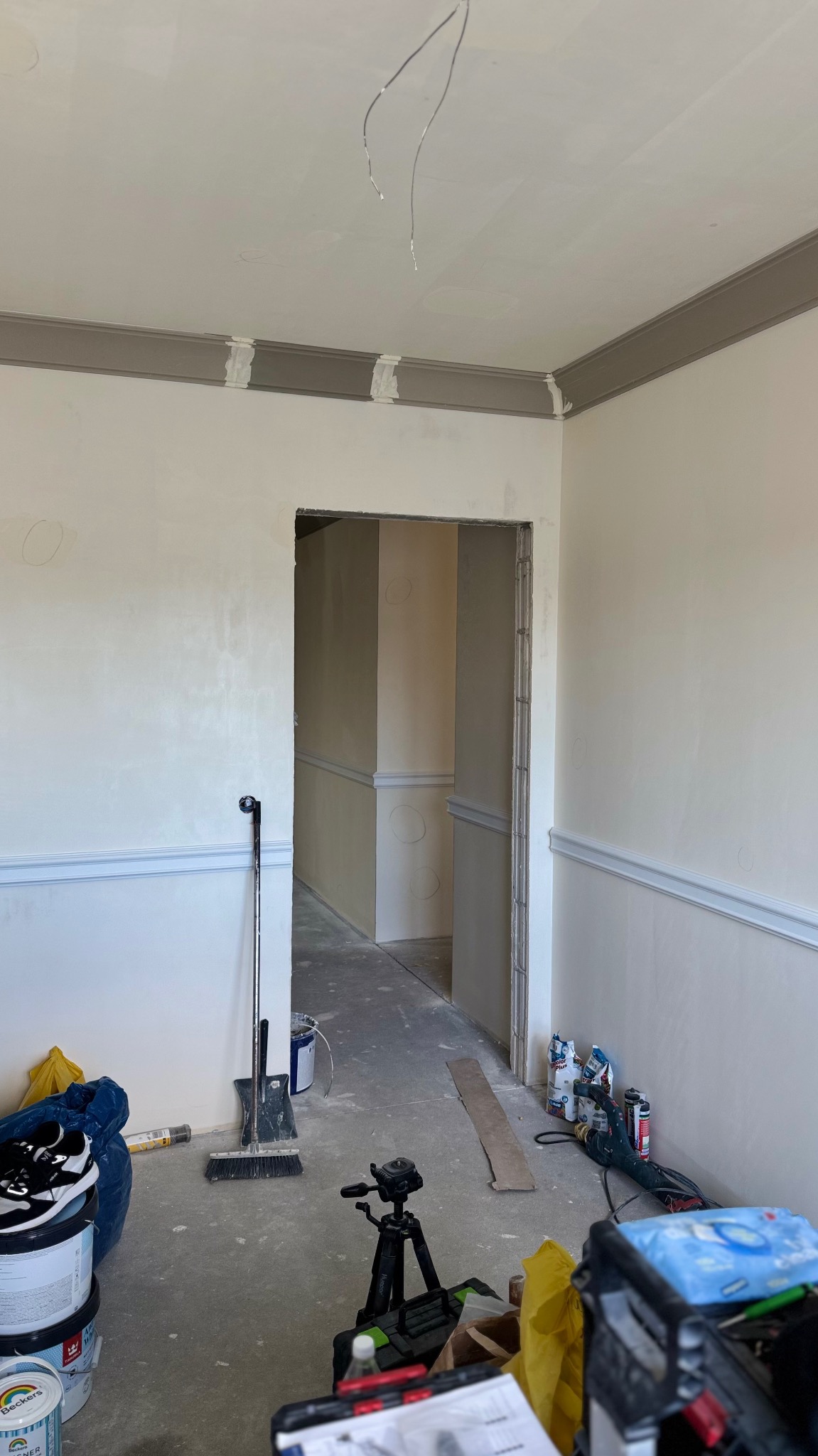 Wnętrze w trakcie prac wykończeniowych: listwy dekoracyjne przy suficie i ścianach, widoczne narzędzia i materiały malarskie, surowy beton na podłodze, przewody wystające z sufitu.