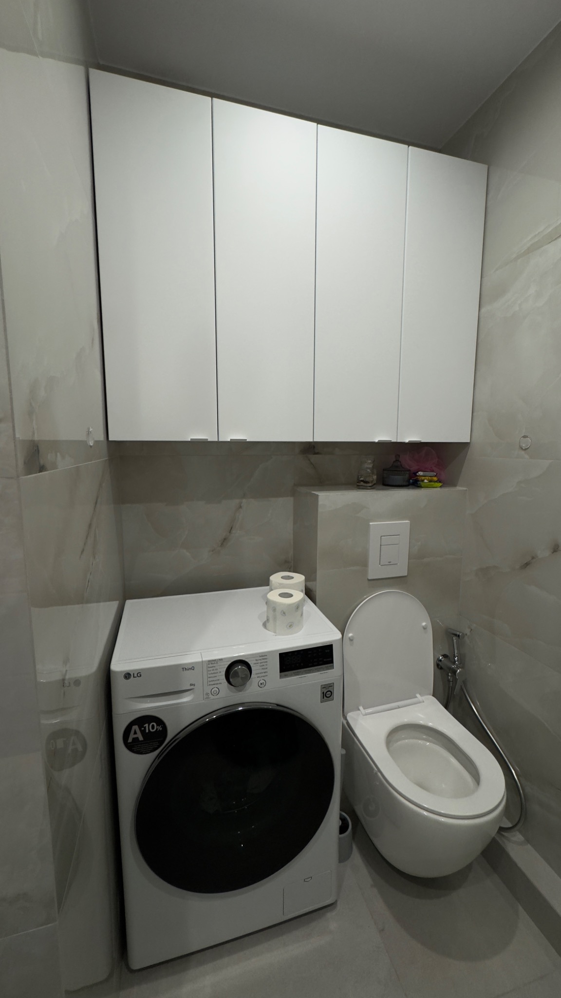 Nowoczesna łazienka z marmurowymi płytkami, białą zabudową meblową, pralką LG i podwieszaną toaletą. Minimalistyczny design, stonowana kolorystyka.