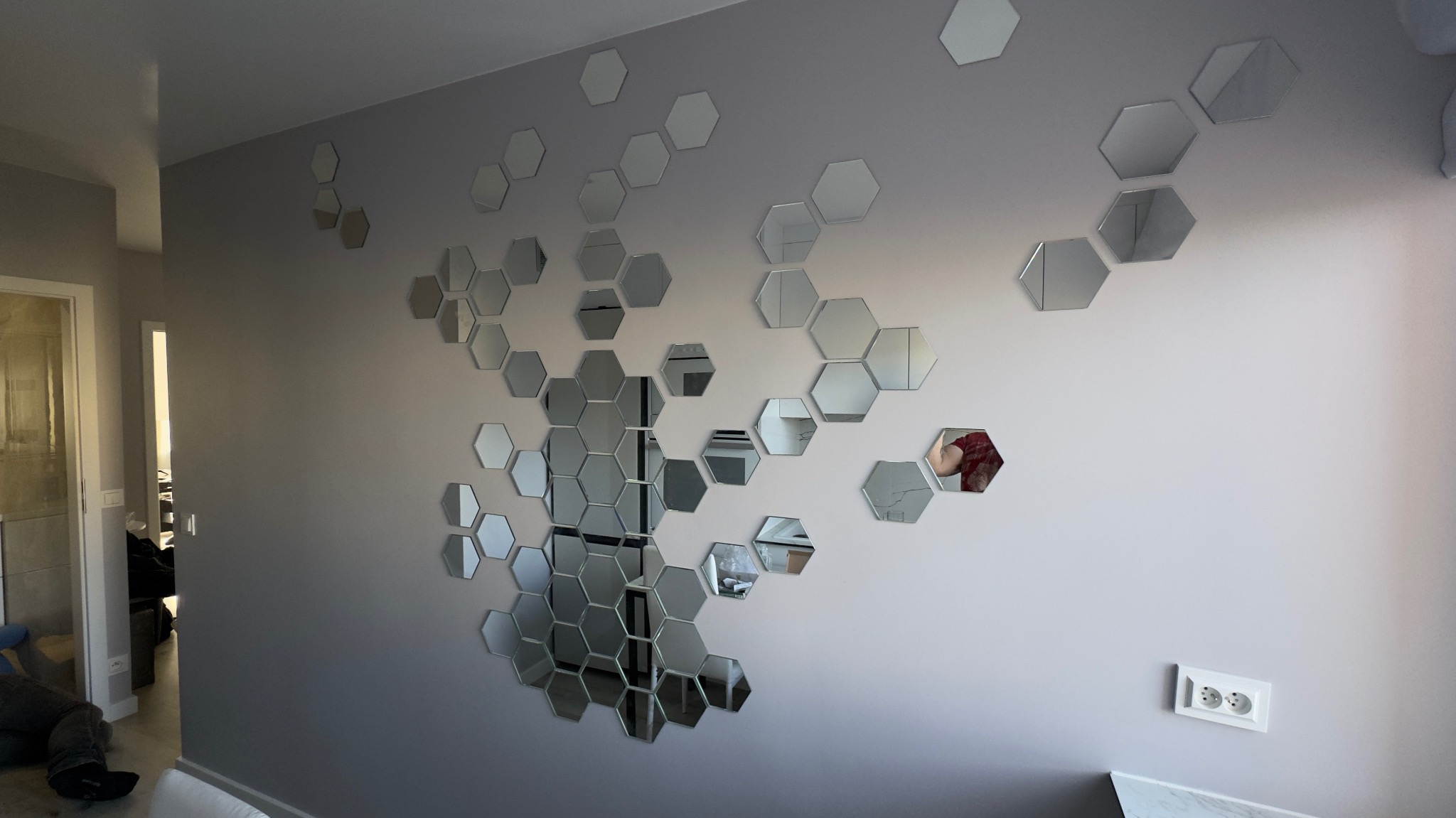 Ściana w pokoju z dekoracyjnymi, lustrzanymi heksagonami tworzącymi wzór plastra miodu. Widoczny fragment gniazdka elektrycznego i odbicie osoby w jednym z luster.
