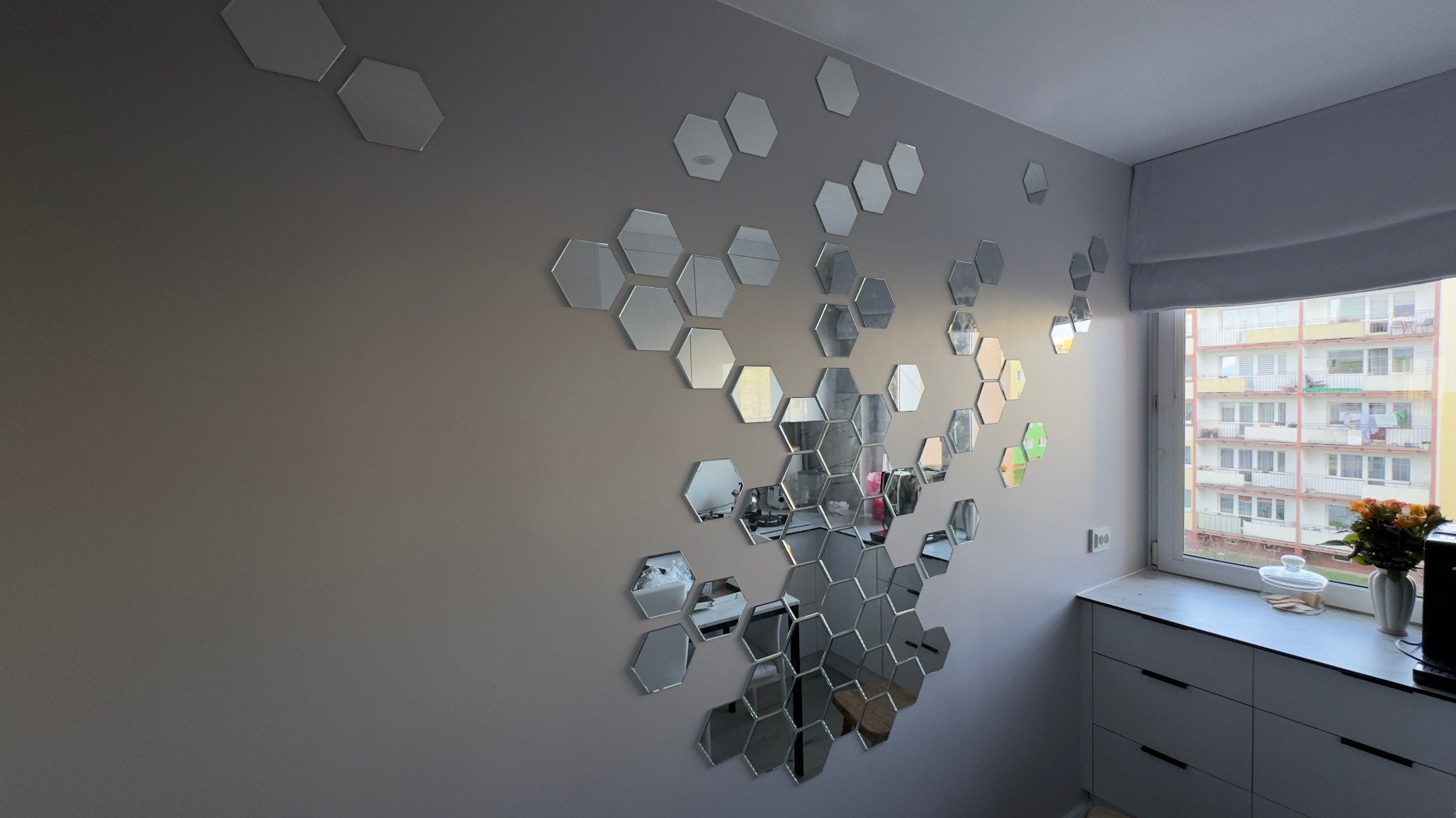 Ściana w pokoju z dekoracją z lustrzanych heksagonów, tworzących abstrakcyjny wzór. W tle okno z widokiem na budynek mieszkalny i fragment blatu kuchennego.