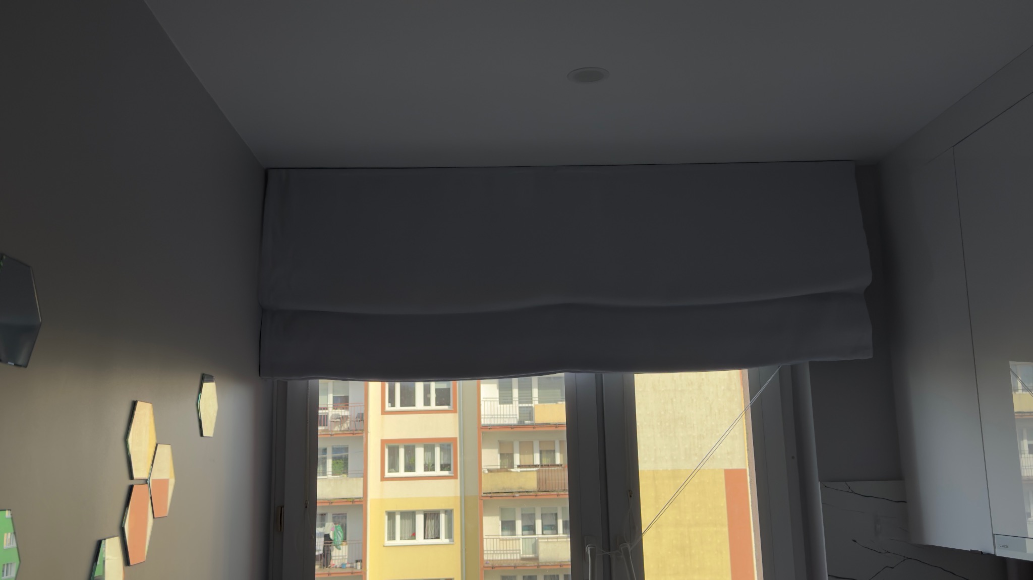 Zamknięta roleta rzymska w jasnym odcieniu, zamontowana w oknie z widokiem na blok mieszkalny. Dekoracyjne, geometryczne elementy na ścianie po lewej stronie.