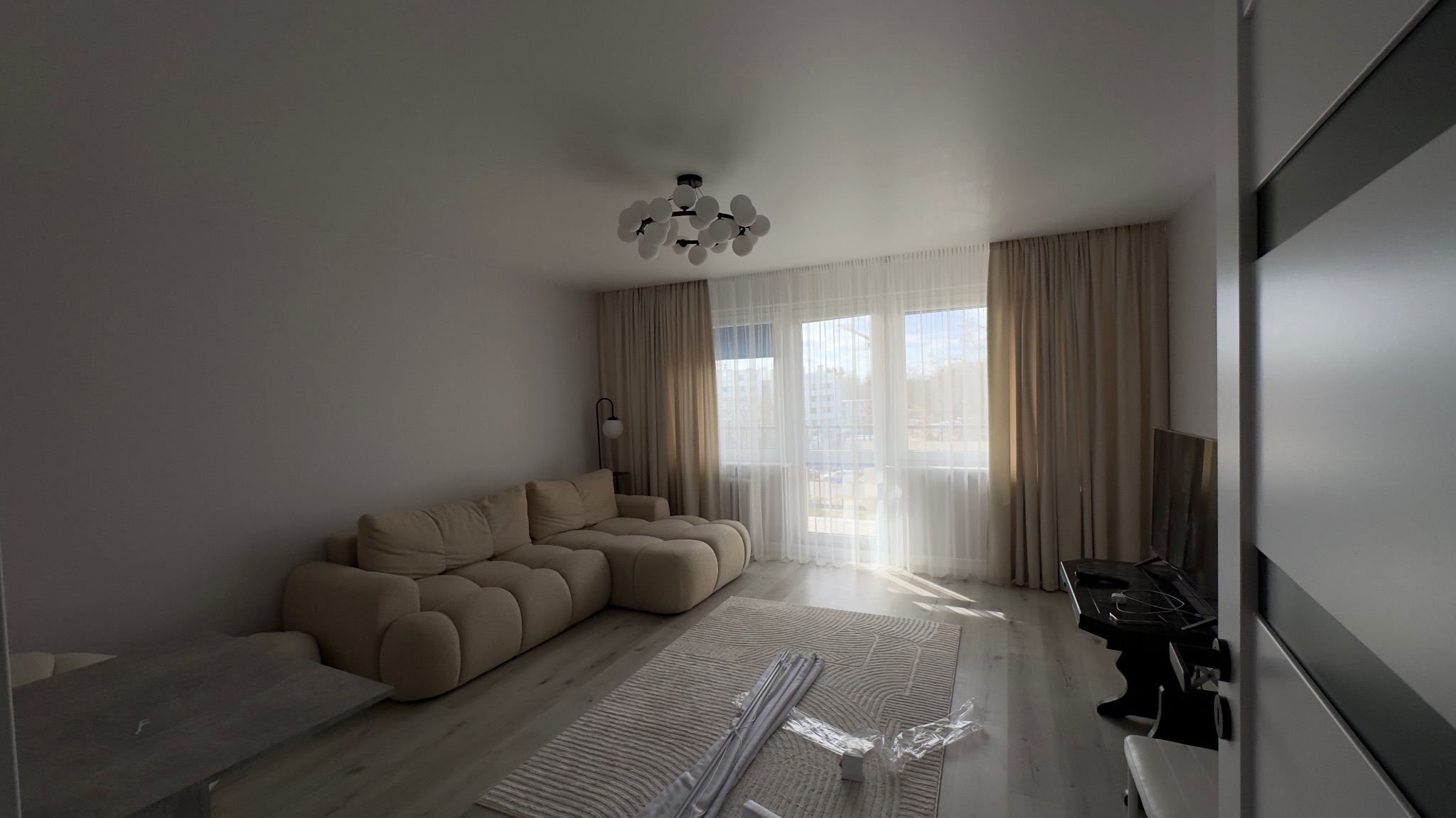 Jasne wnętrze salonu z beżową sofą modułową, wzorzystym dywanem i nowoczesną lampą sufitową. Okno z firanami wpuszcza naturalne światło. Minimalistyczny wystrój.