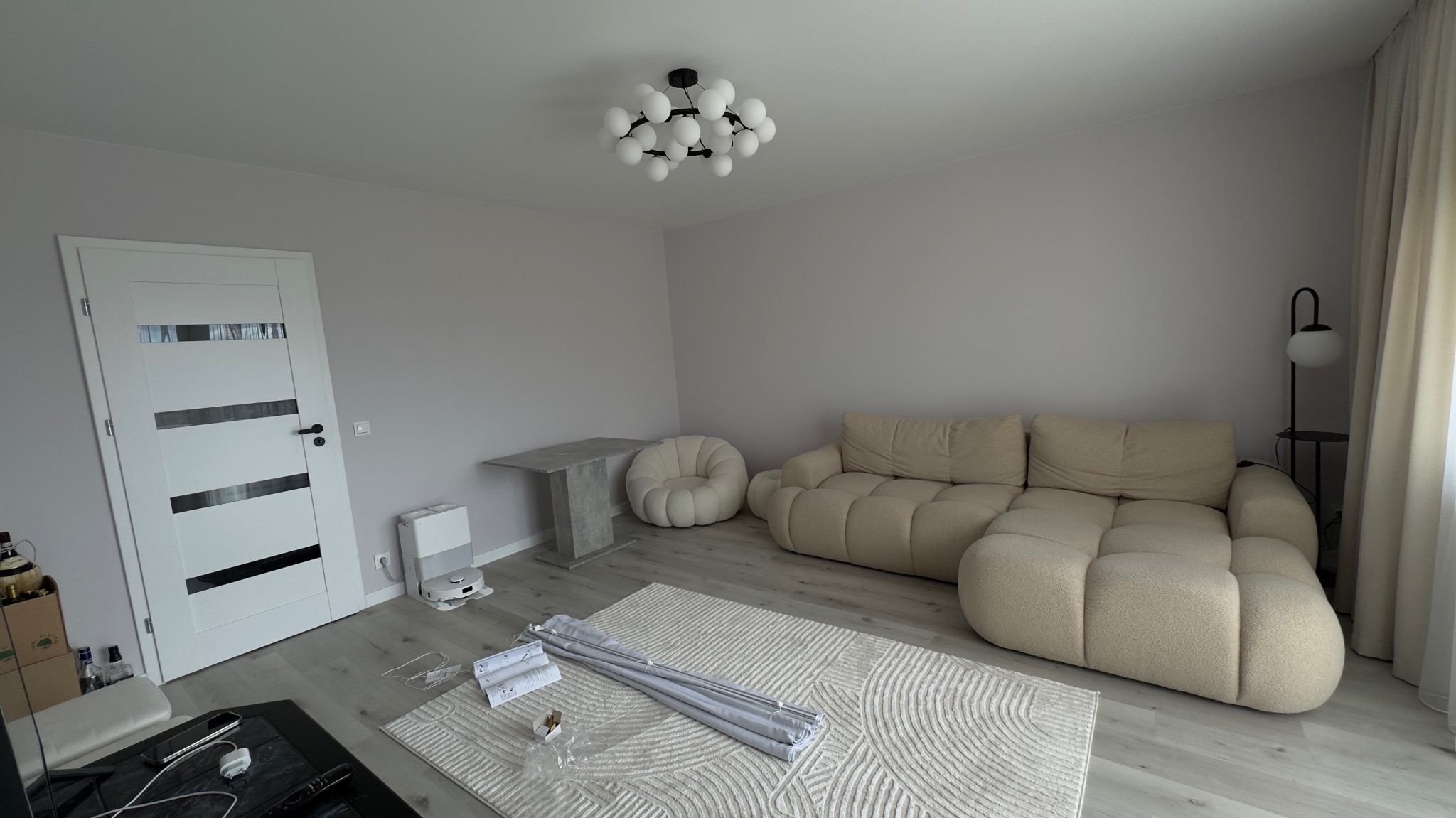 Jasne wnętrze pokoju dziennego z beżową sofą modułową, stolikiem, pufą i lampą. Na podłodze leżą elementy do montażu. Widoczna biała ściana i drzwi.