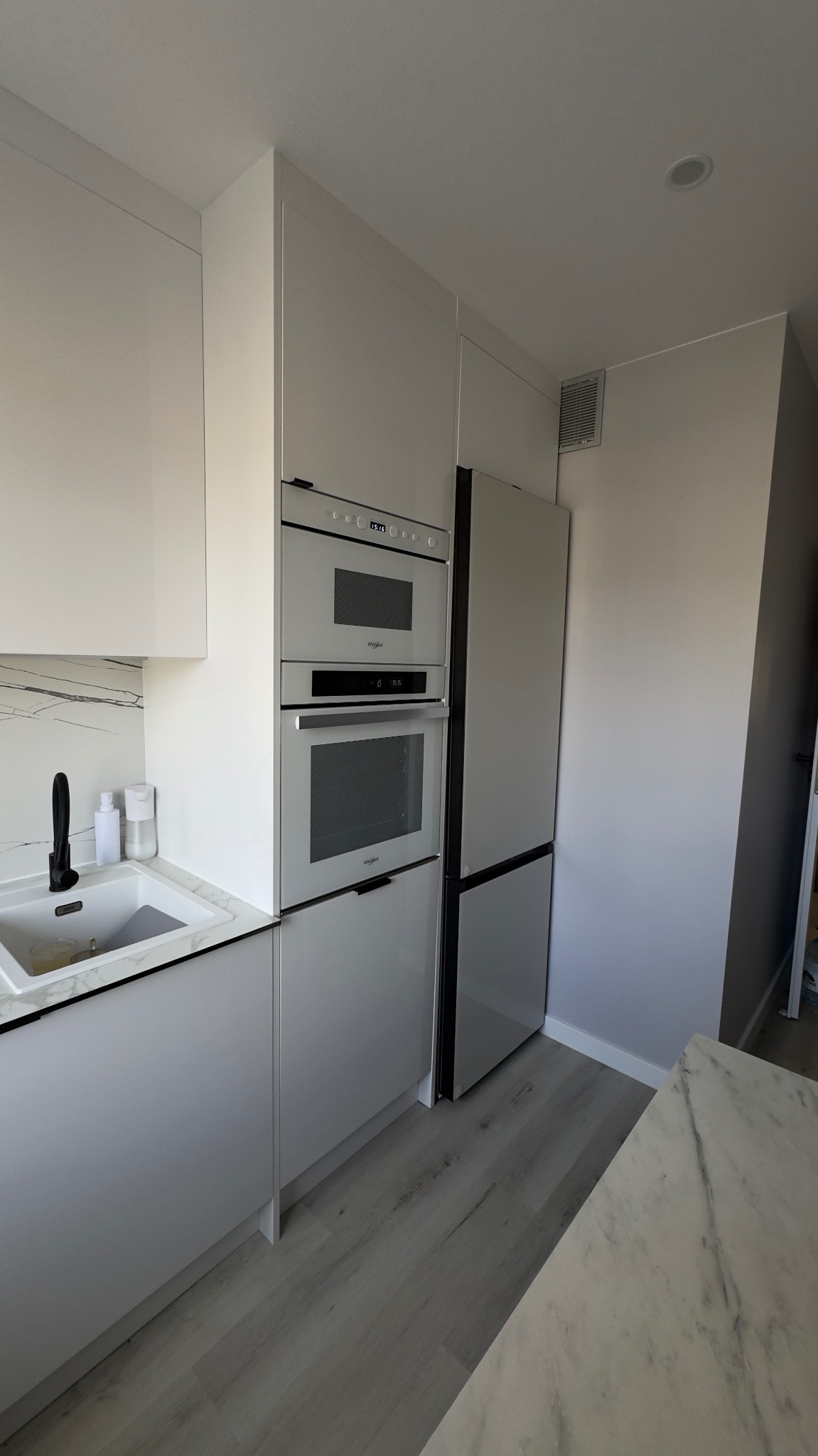 Nowoczesna, jasna kuchnia z zabudową w odcieniach szarości i bieli. Widoczny zlew z czarną armaturą, zabudowane sprzęty AGD i fragment blatu imitującego marmur.