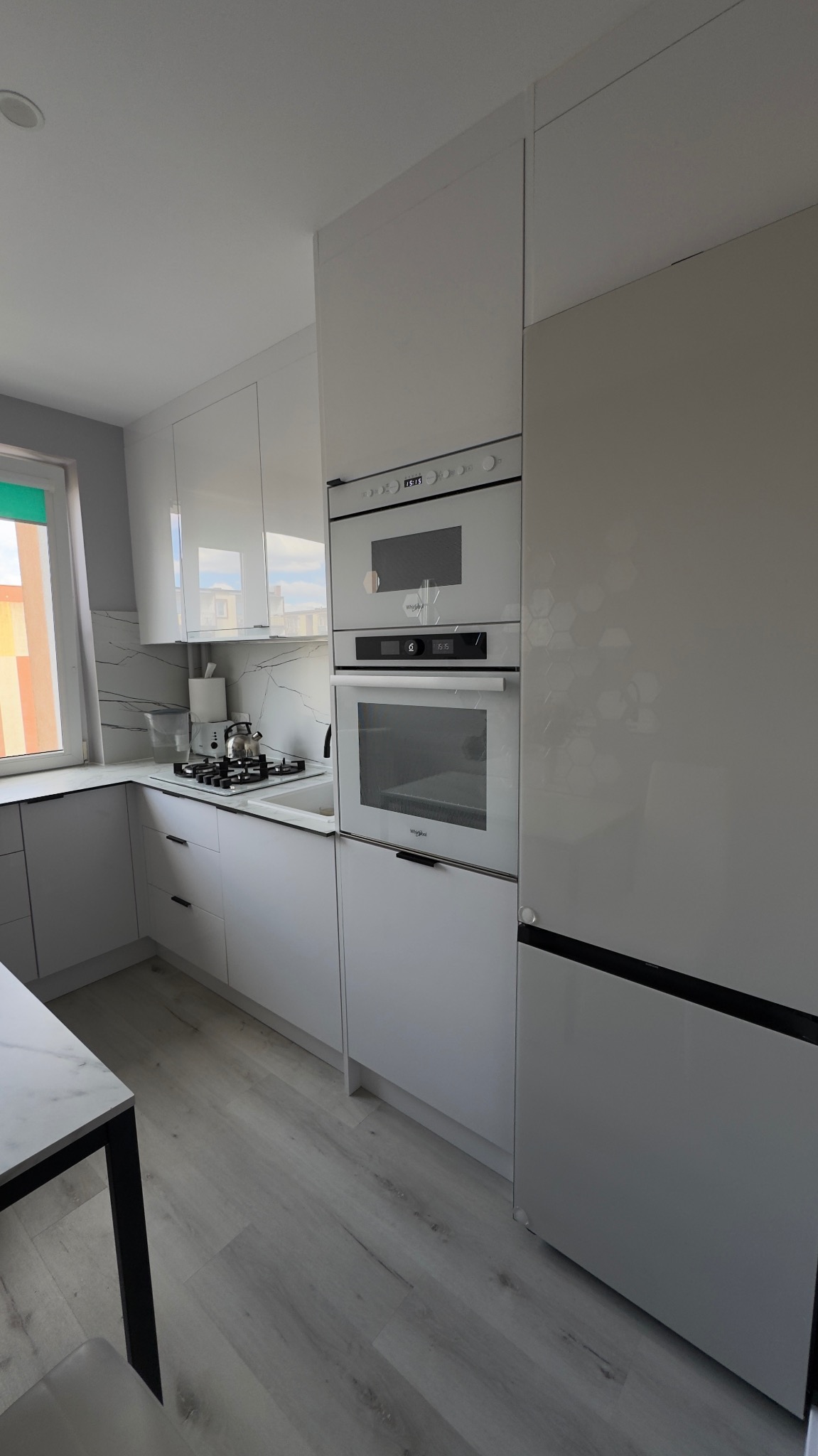 Nowoczesna, jasna kuchnia z białymi, błyszczącymi frontami szafek i sprzętami AGD w zabudowie. Marmurowy wzór na ścianie nad blatem. Widok na okno.