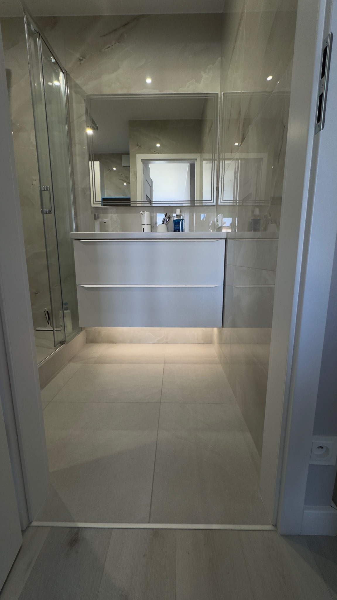 Nowoczesna łazienka z podświetlaną szafką pod umywalką, kabiną prysznicową i lustrem. Jasne płytki podłogowe i marmurowe wykończenie ścian.