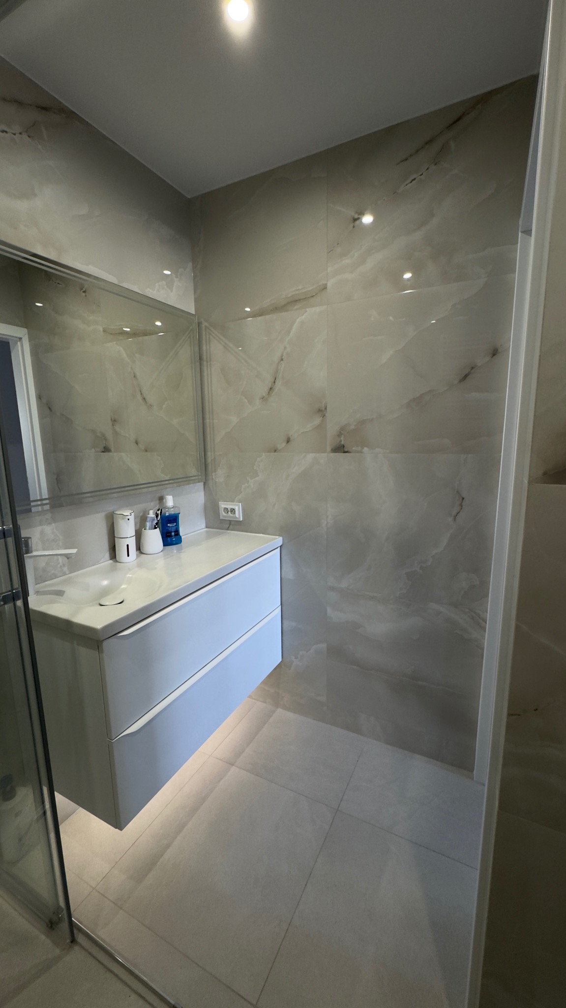 Nowoczesna łazienka z marmurowymi płytkami, podwieszaną szafką z umywalką i lustrem. Minimalistyczny design, jasne kolory i subtelne oświetlenie.