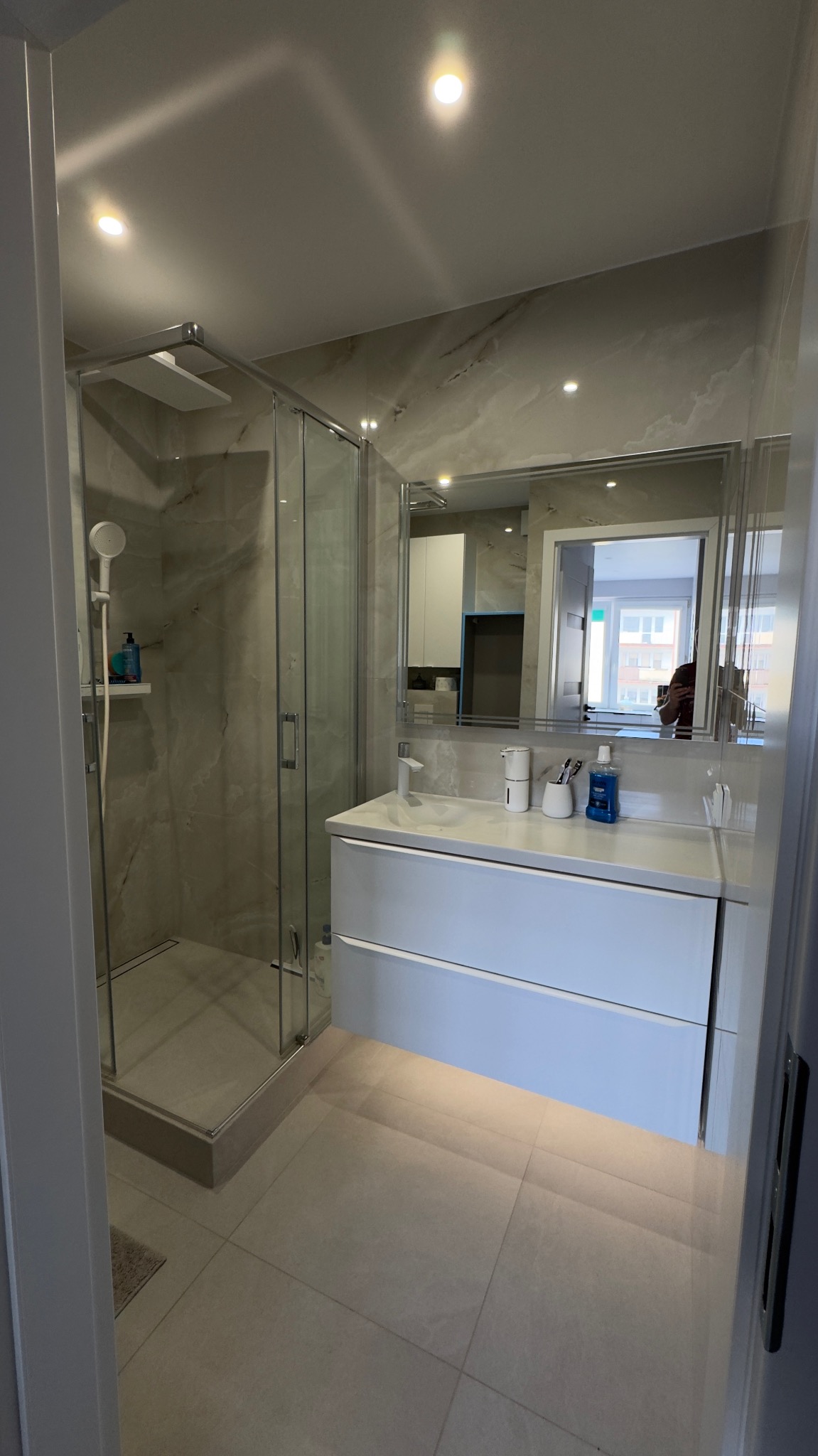 Nowoczesna łazienka z kabiną prysznicową, umywalką z szafką i dużym lustrem. Jasne płytki imitujące marmur na ścianach i podłodze. Oświetlenie punktowe.