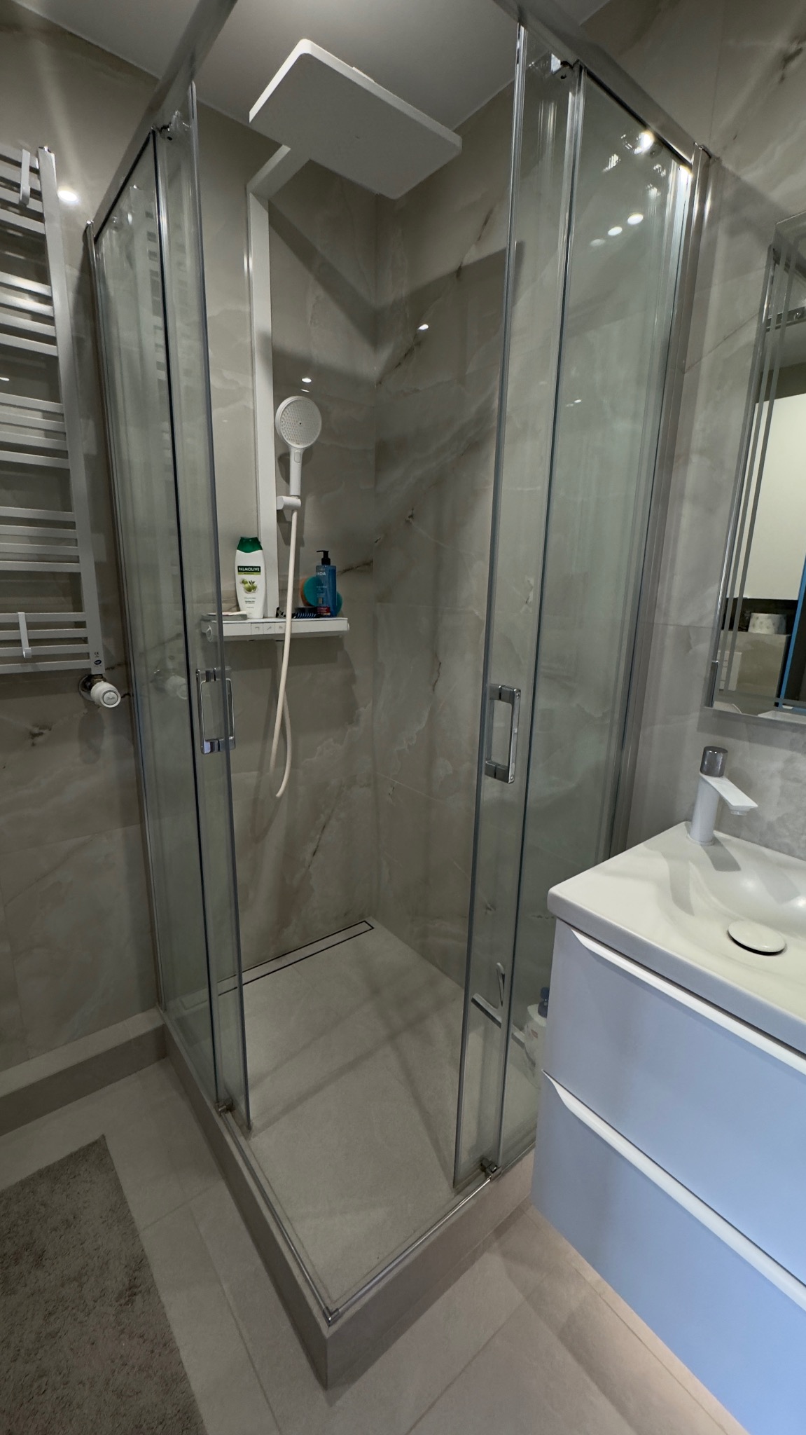 Nowoczesna kabina prysznicowa z deszczownicą i odpływem liniowym, wykończona płytkami imitującymi marmur. Biała umywalka z szafką w tle. Minimalistyczny design.