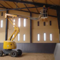 Żółty podnośnik koszowy z pracownikiem w kasku, wykonującym prace na wysokości wewnątrz hali z beżowymi ścianami i rzędami okien.