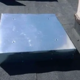 Metalowy właz dachowy, kwadratowy, z czterema otworami na wierzchu, zamontowany na czarnej papie. Widoczny komin w tle.