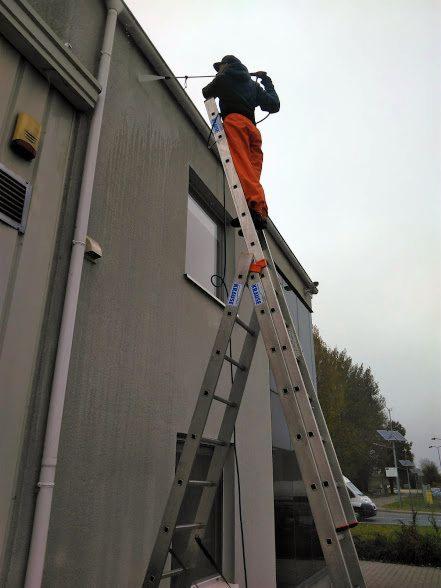 Pracownik w pomarańczowych spodniach na aluminiowej drabinie, spryskuje elewację budynku myjką ciśnieniową, widoczne ślady po umyciu.