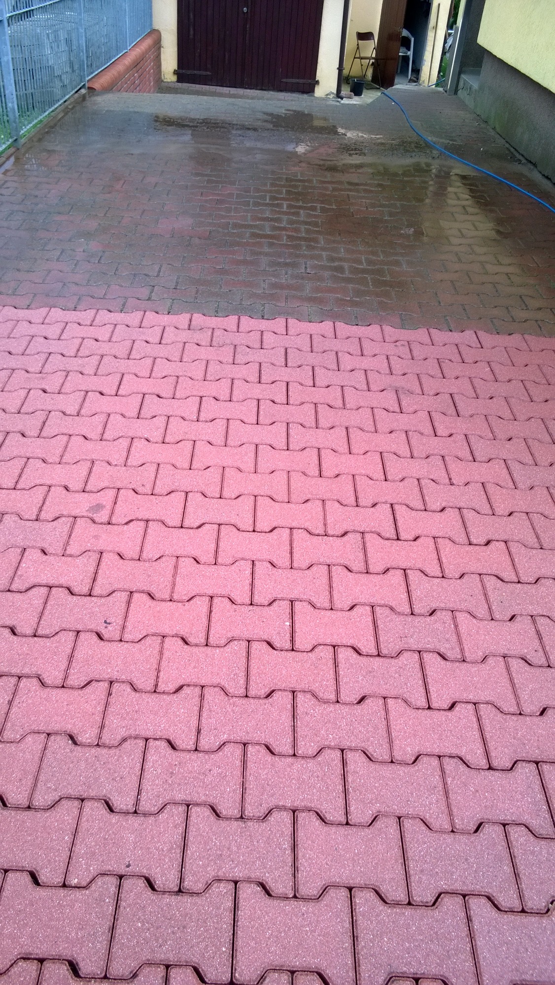 Częściowo umyta kostka brukowa w kolorze czerwonym, widoczna różnica w czystości przed i po myciu, w tle brama garażowa i fragment budynku.