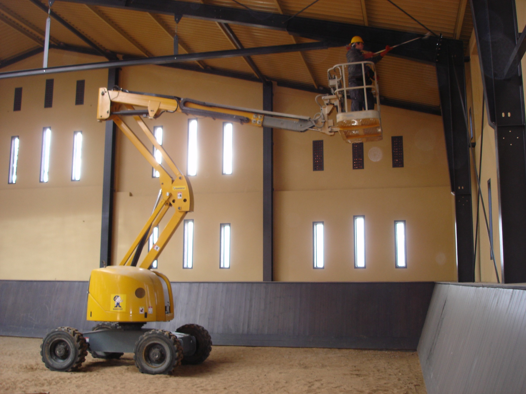 Żółty podnośnik koszowy z pracownikiem w kasku, wykonującym prace na wysokości wewnątrz hali z beżowymi ścianami i rzędami okien.