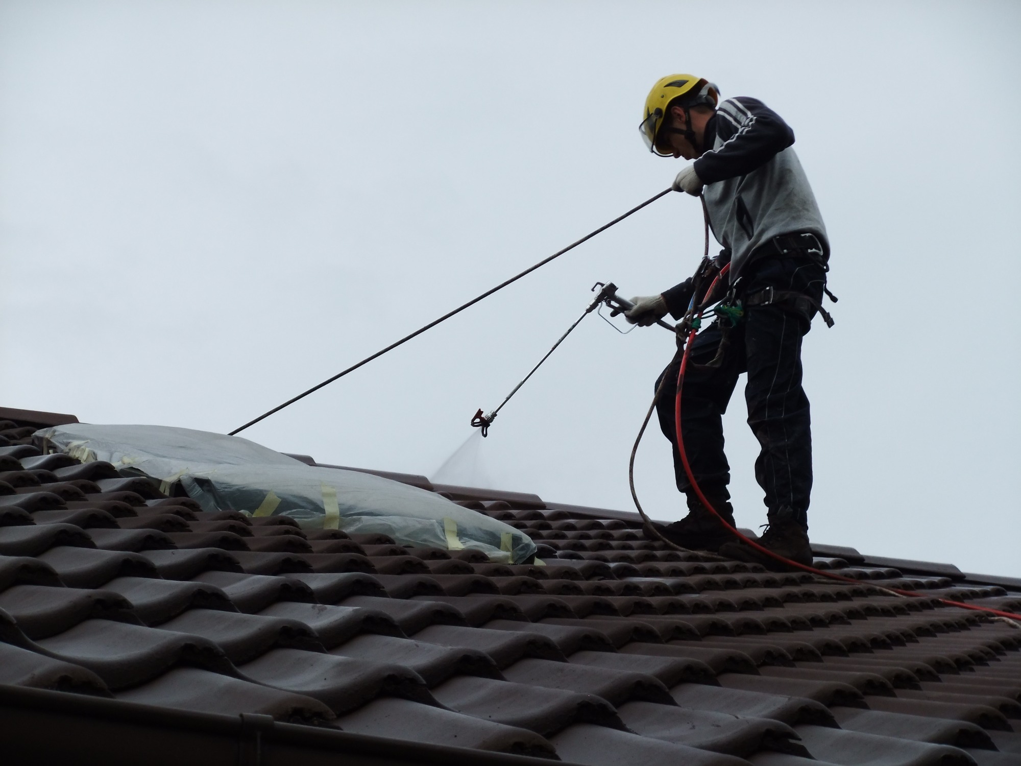 Pracownik w kasku, zabezpieczony linami, aplikuje preparat na dach w Szczecinie za pomocą specjalistycznego pistoletu natryskowego. Część dachu zabezpieczona folią.