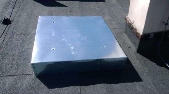 Metalowy właz dachowy, kwadratowy, z czterema otworami na wierzchu, zamontowany na czarnej papie. Widoczny komin w tle.