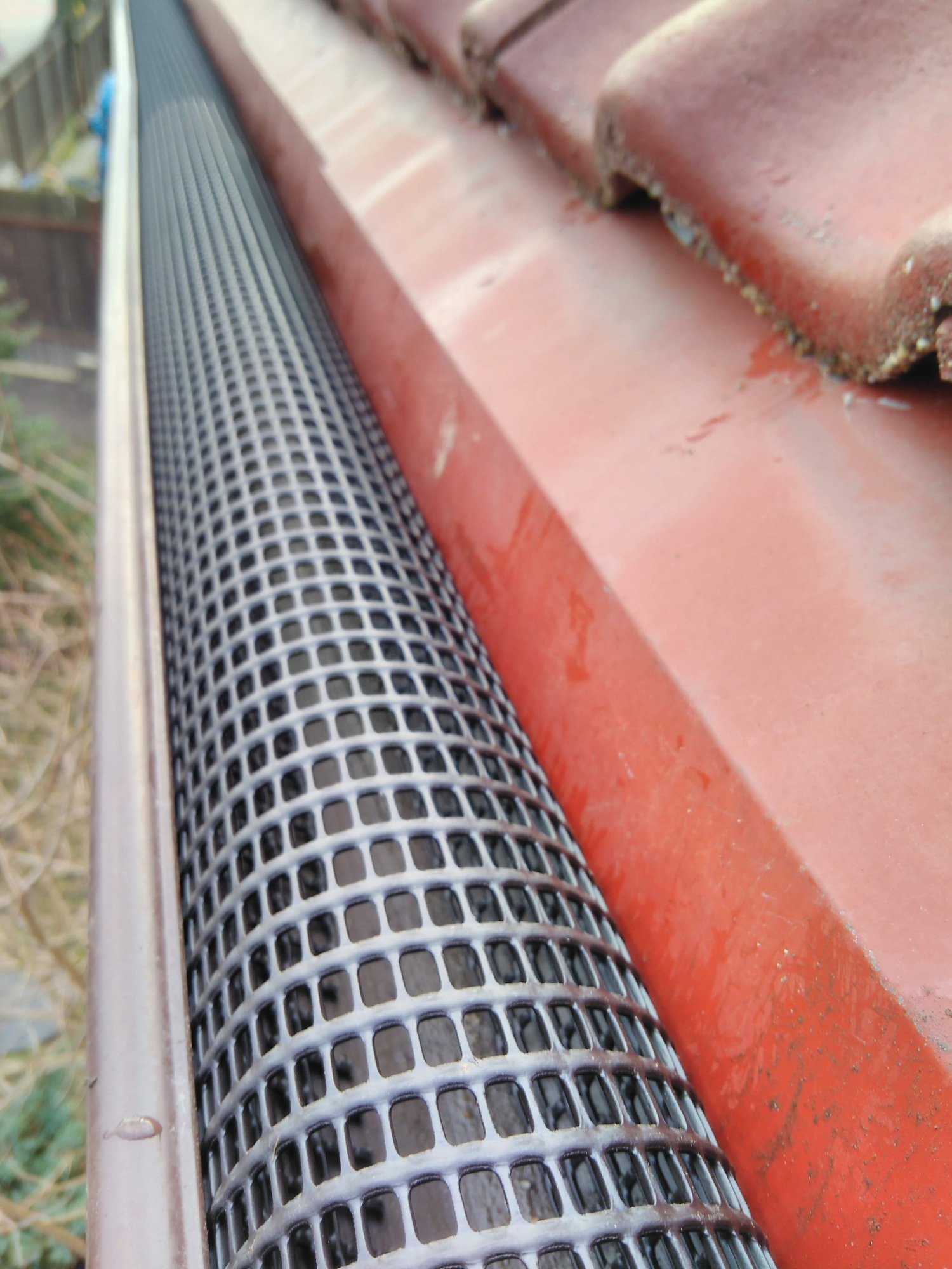 Zamontowana siatka ochronna w rynnie, zabezpieczająca przed liśćmi, widok z bliska na czerwony dach z dachówki.