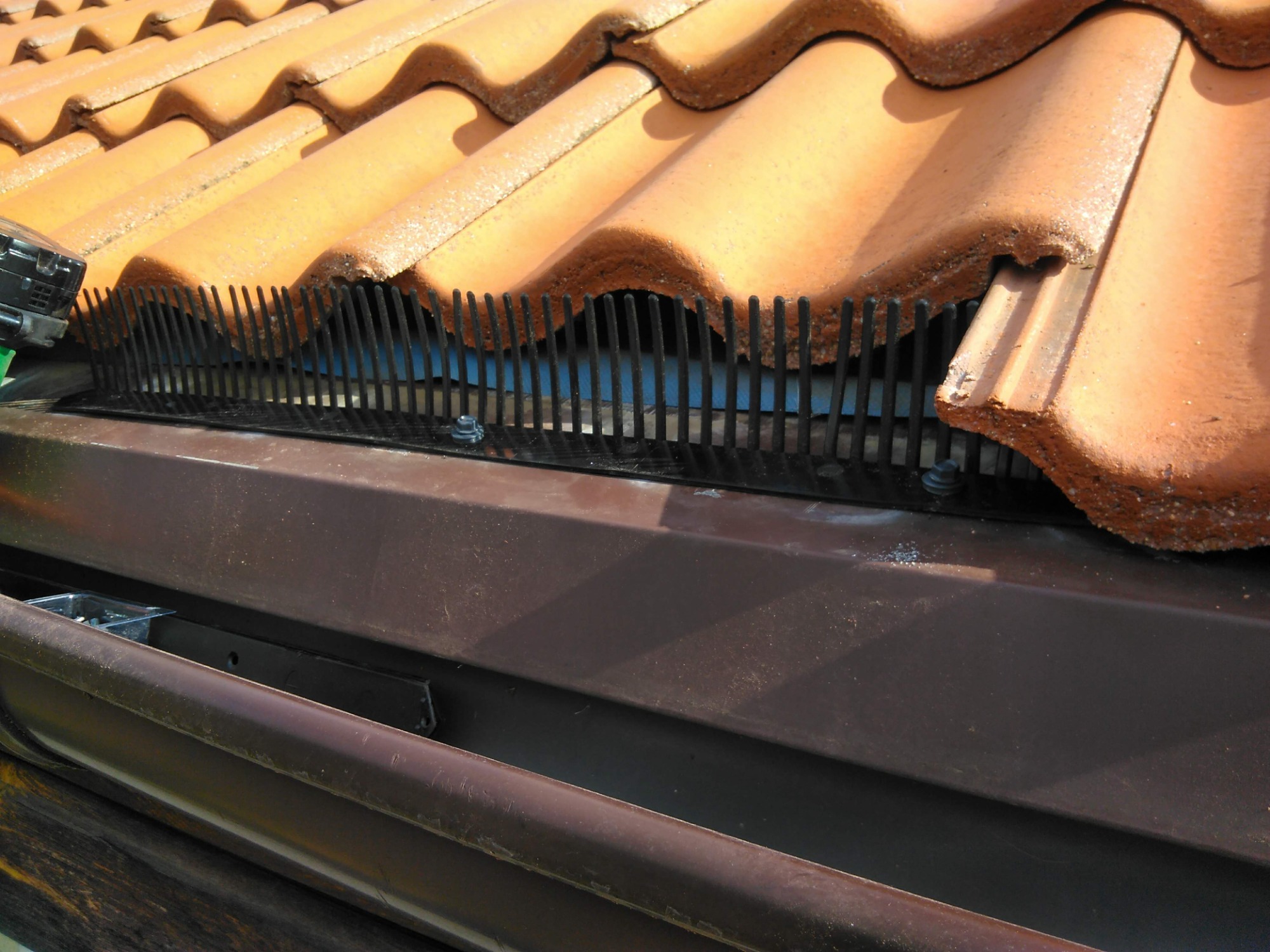 Zamontowana wróblówka pod dachówką ceramiczną, widoczna brązowa rynna i elementy mocujące.