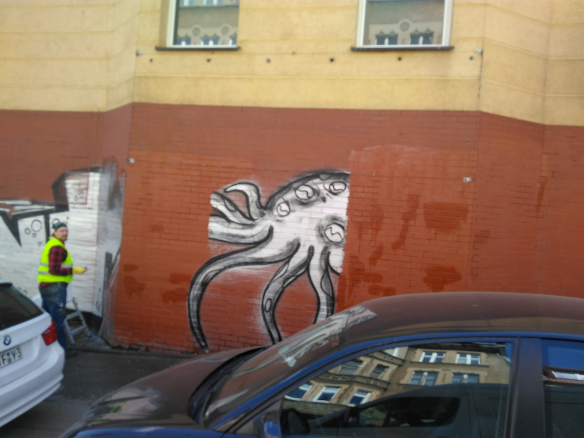 Mężczyzna w odblaskowej kamizelce usuwa graffiti przedstawiające ośmiornicę z klinkierowej ściany budynku, widoczne ślady mycia i częściowo usunięte graffiti po lewej stronie.