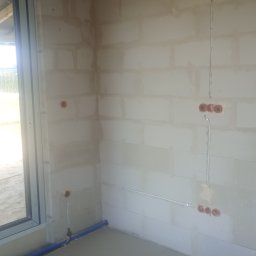 ZWARCIE Mateusz Mioduszewski - Surowy pokój z instalacją elektryczną w ścianach z betonu komórkowego. Widoczne puszki pod gniazdka i kable. W tle okno i fragment podłogi z rurą.