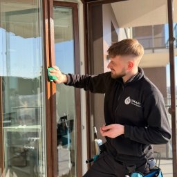 SmartCleaning - Uśmiechnięty mężczyzna w czarnej bluzie z logo Smart Cleaning myje okno na balkonie, używając zielonej gąbki. Widoczne narzędzia w niebieskiej torbie.