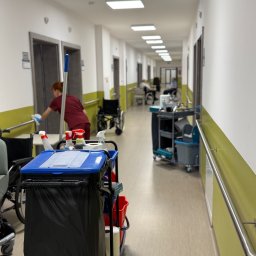 SmartCleaning - Wózek serwisowy z chemią i mopem na korytarzu w Warszawie. W tle osoba w uniformie czyści poręcz. Widoczne wózki inwalidzkie i osoby na wózkach.
