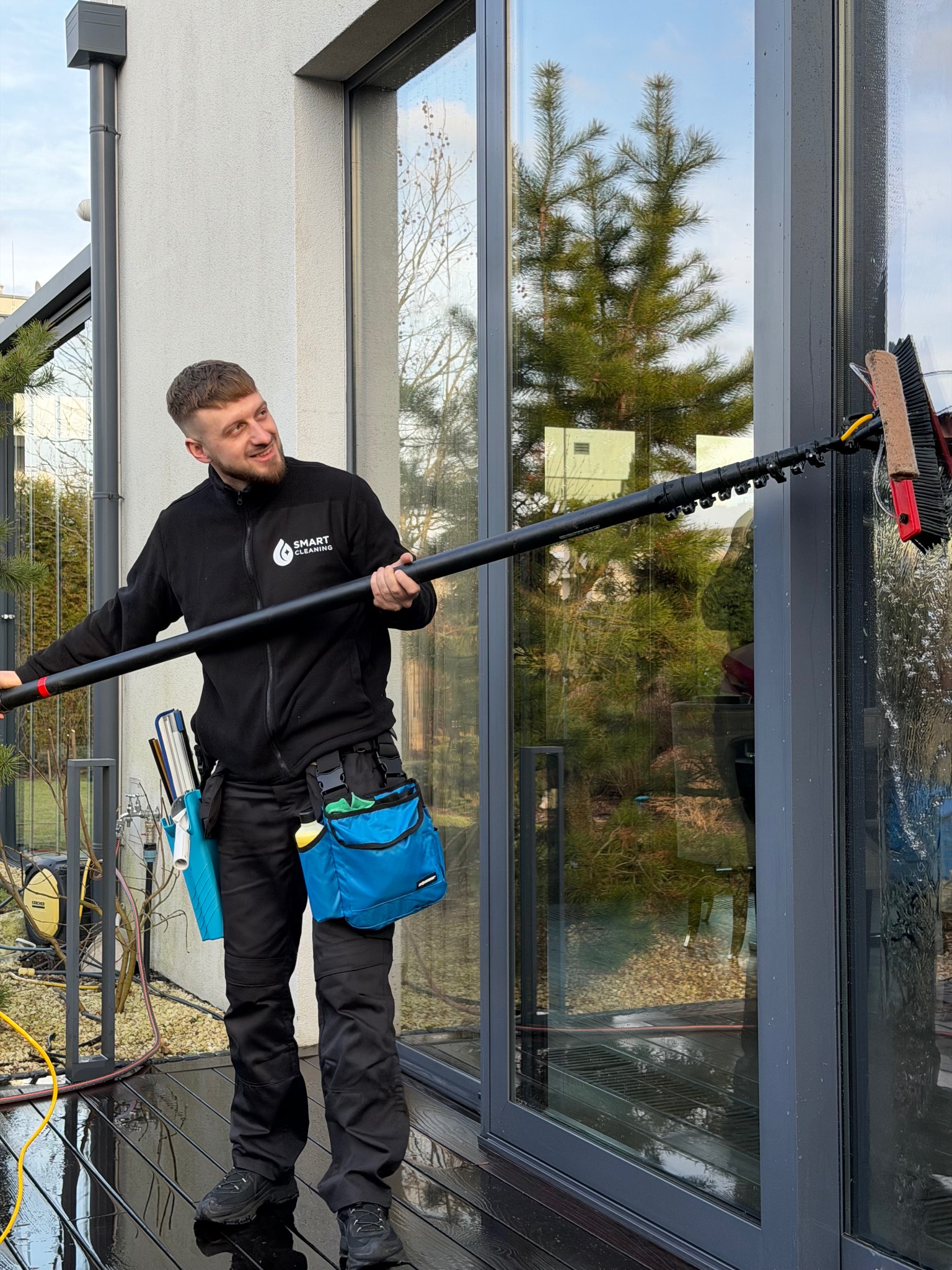 Pracownik w czarnym uniformie z logo firmy czyści okno na zewnątrz budynku za pomocą teleskopowego kija z szczotką. Widoczne narzędzia i akcesoria.