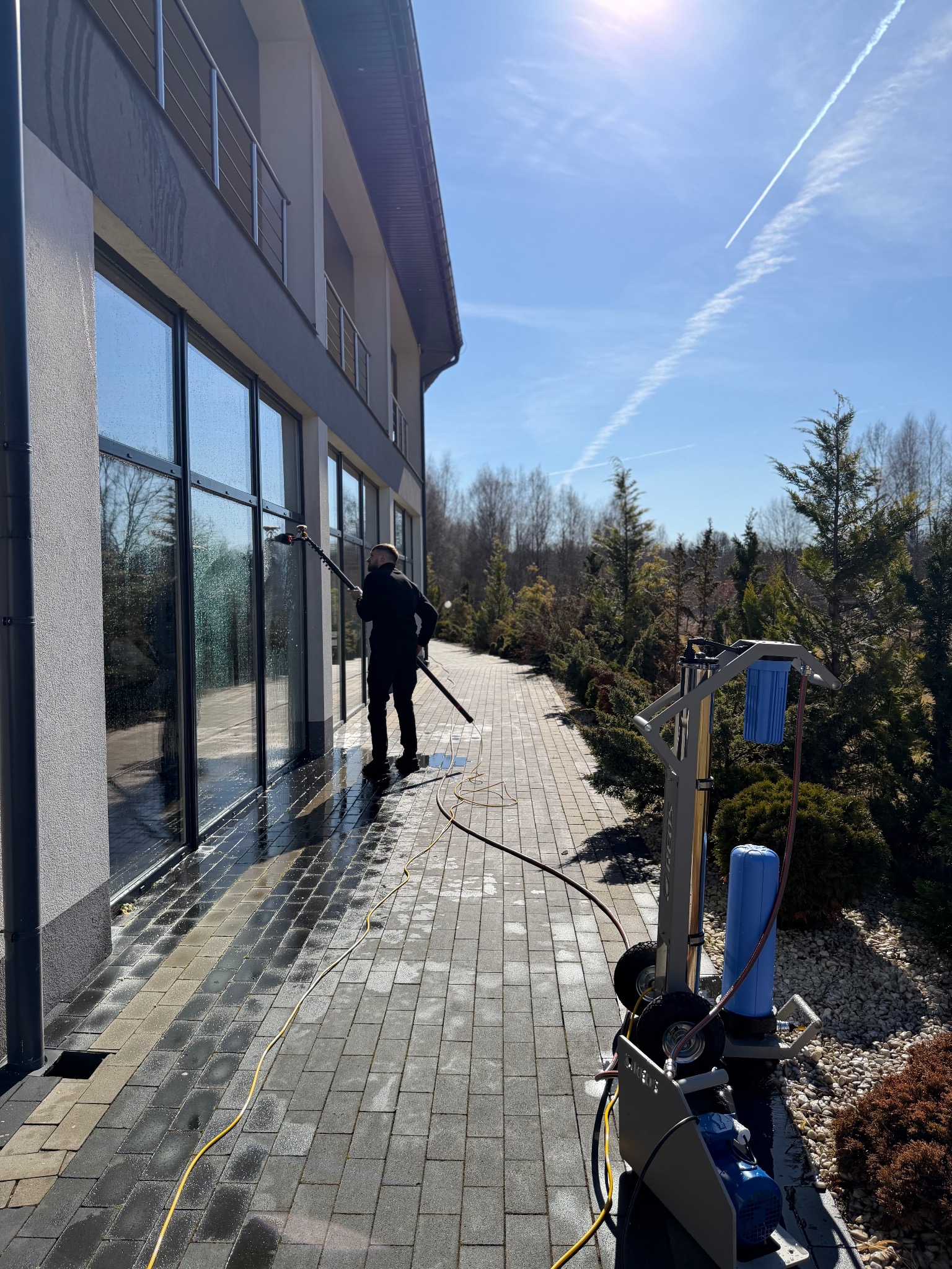 Pracownik w czarnym kombinezonie czyści okna budynku za pomocą specjalistycznego sprzętu. Widoczny wózek z filtrami i wąż doprowadzający wodę. Słoneczny dzień.