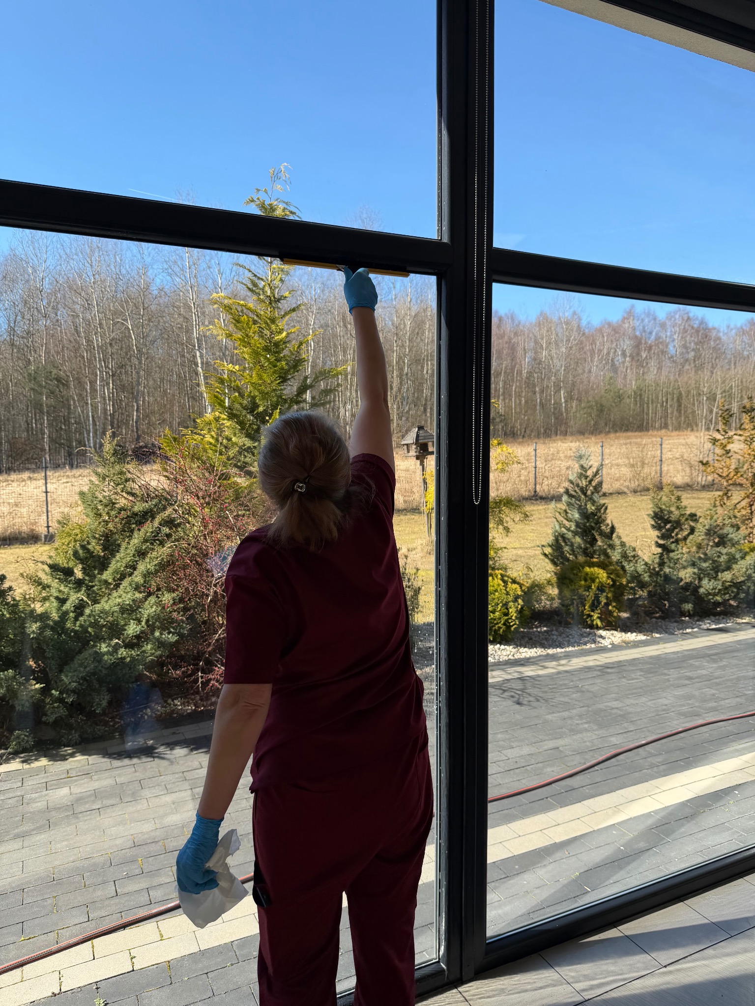 Osoba w bordowym uniformie i rękawicach czyści okno panoramiczne w domu. Widok na ogród z zielenią i drzewami w tle. Słoneczny dzień, czyste niebo.