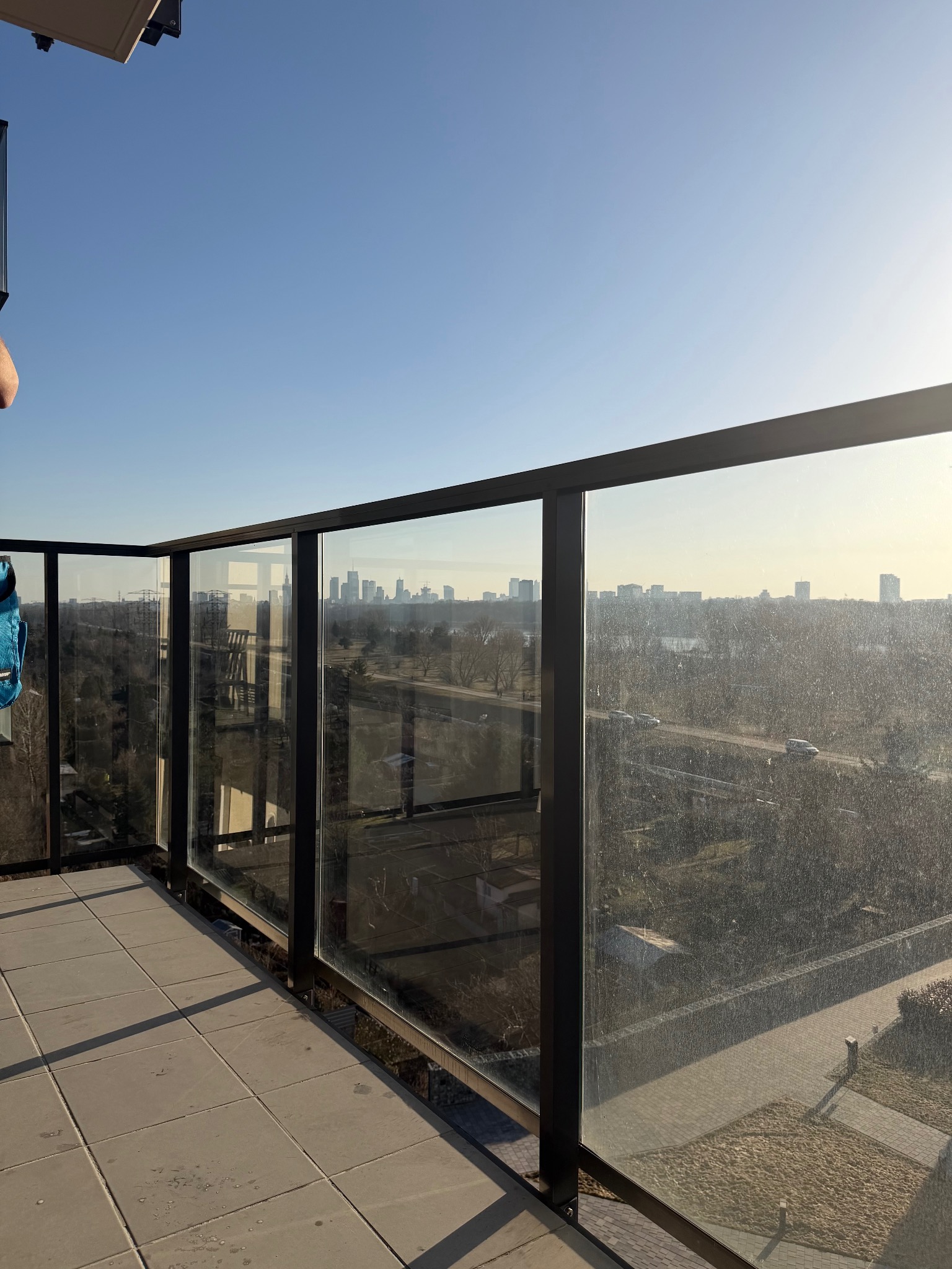 Balkon z brudnymi szybami i widokiem na miasto w oddali. Słoneczny dzień, widoczne zacieki na szkle. Perspektywa z góry, szare płytki na podłodze balkonu.