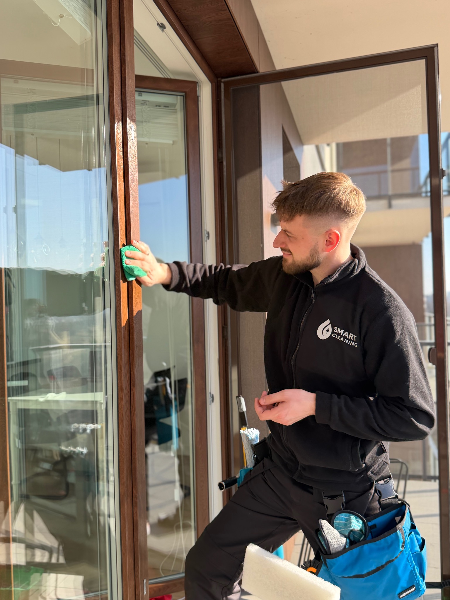 Uśmiechnięty mężczyzna w czarnej bluzie z logo Smart Cleaning myje okno na balkonie, używając zielonej gąbki. Widoczne narzędzia w niebieskiej torbie.
