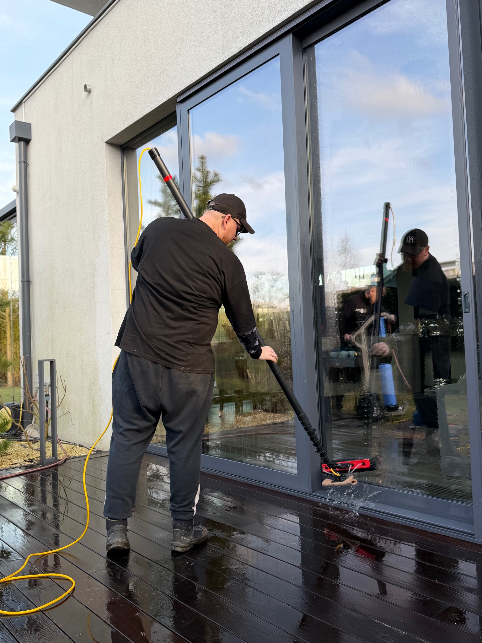 Mężczyzna w czarnym ubraniu czyści okno na zewnątrz domu za pomocą specjalistycznego sprzętu. W tle widoczny drugi pracownik i odbicie nieba w szybie.