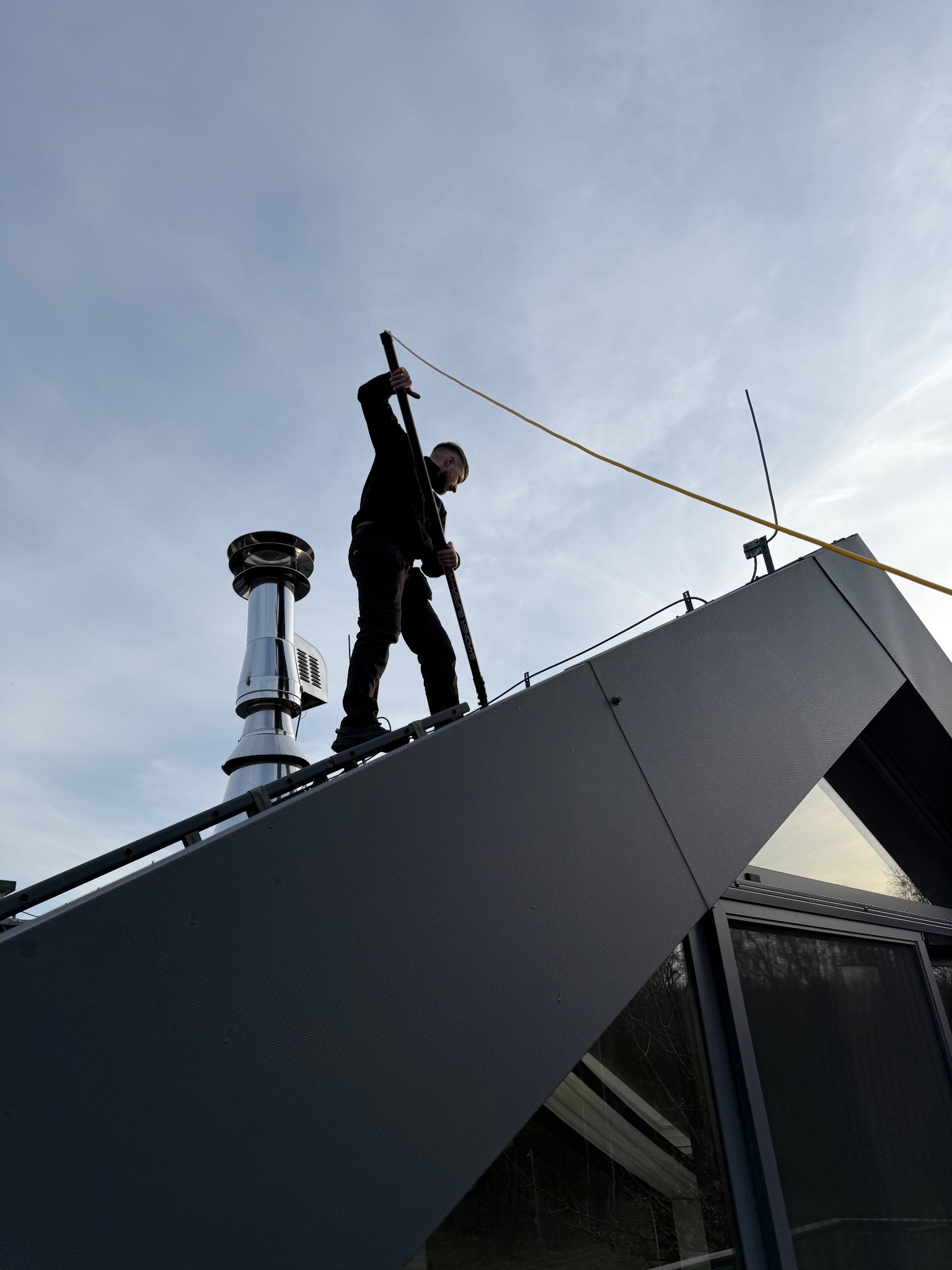 Pracownik na dachu czyści komin nowoczesnego domu z użyciem długiej szczotki. Widok z dołu na tle jasnego nieba.