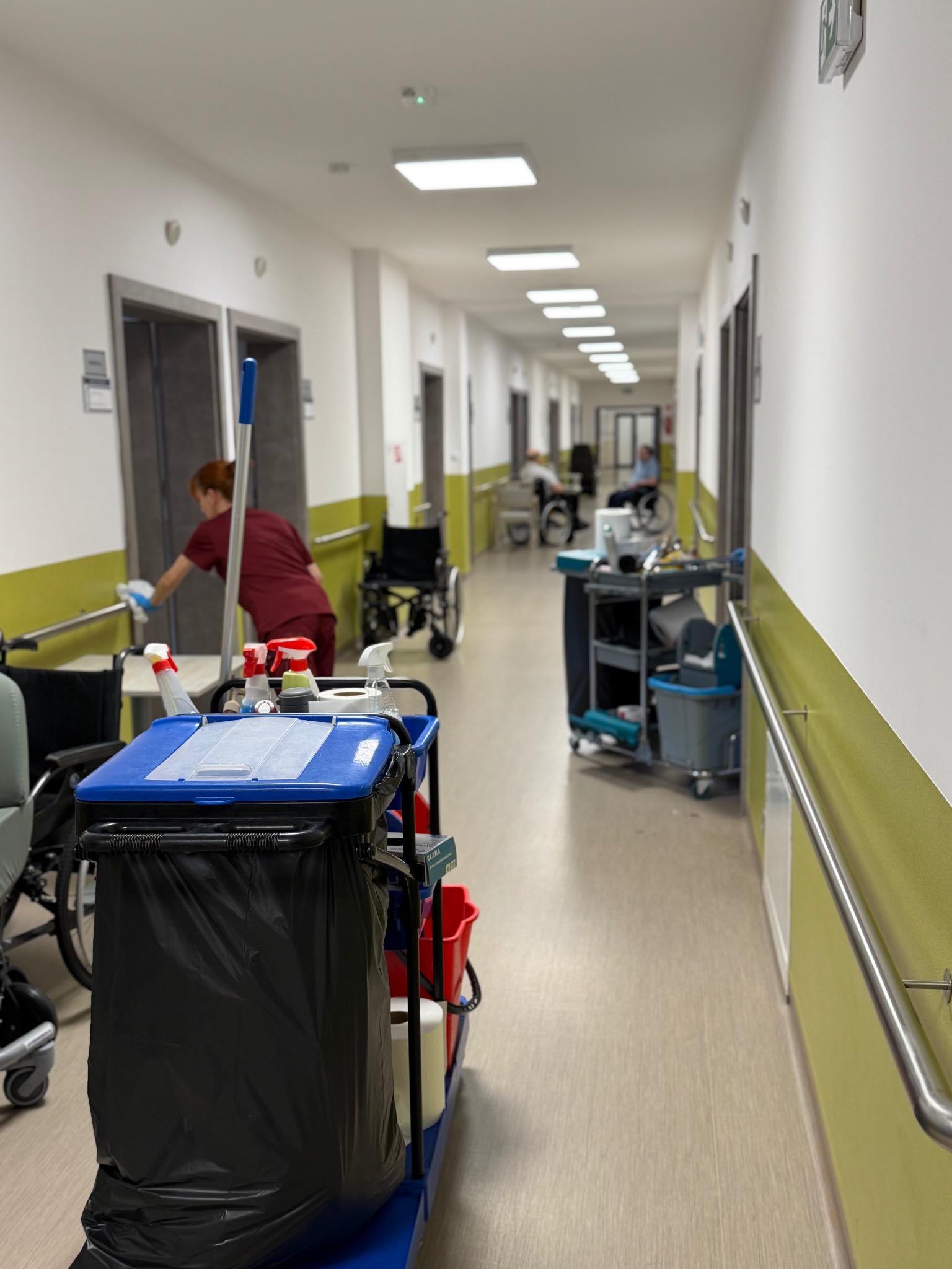 Wózek serwisowy z chemią i mopem na korytarzu w Warszawie. W tle osoba w uniformie czyści poręcz. Widoczne wózki inwalidzkie i osoby na wózkach.