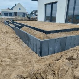 DAMI-BRUK Usługi Brukarskie Daniel Sobota - Przygotowanie podłoża pod taras z obrzeżami z betonowych płyt. W tle dom z dużymi oknami i ekipa budowlana. Piasek, ziemia i elementy konstrukcyjne.