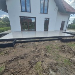 DAMI-BRUK Usługi Brukarskie Daniel Sobota - Wykończony taras z jasnych płytek przy białym domu z brązowymi oknami. Widoczne czarne obramowanie tarasu i przygotowany teren wokół. Prace w toku.