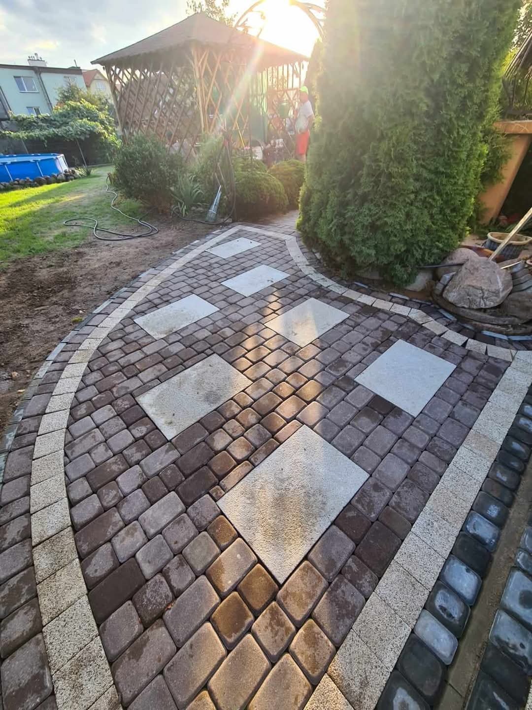 Zakrzywiony chodnik z kostki brukowej w odcieniach brązu i beżu, z kwadratowymi płytami. W tle altana, trawnik i basen. Słoneczne oświetlenie.